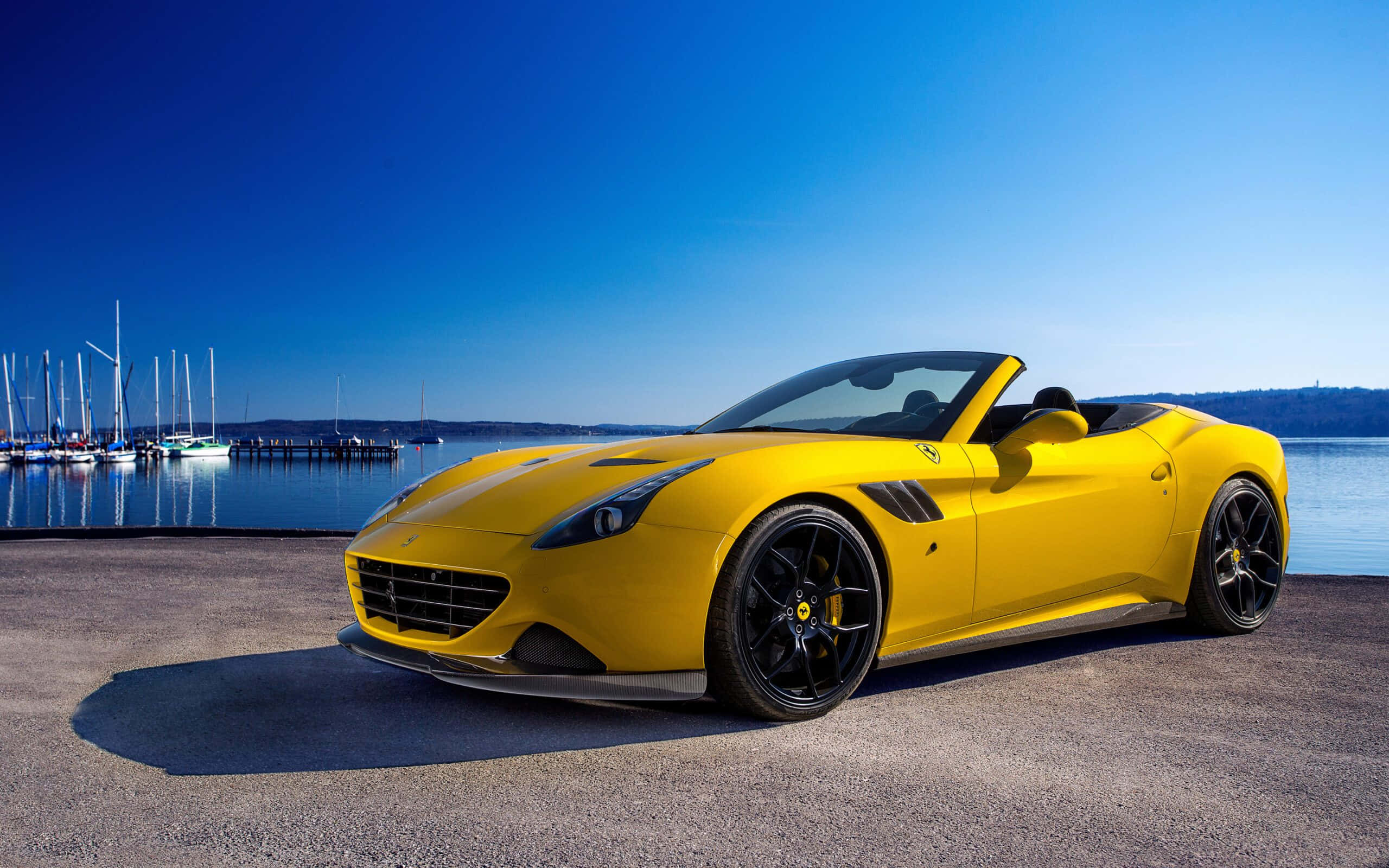 Ferrari California T 桌布 壁紙 — 一輛鮮黃色的法拉利敞篷車停泊在碼頭上，背景有帆船和寧靜的水面，天空清澈湛藍。