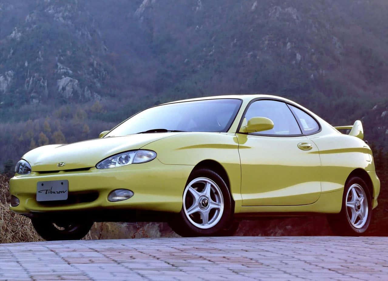 Hyundai Tiburon 桌布 壁紙 — 一輛鮮黃色的現代 Tiburon 雙門跑車停放在石徑上，背景是岩石山脈和秋天的樹木。