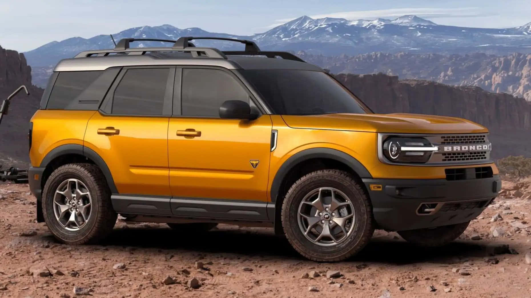Ford Bronco 桌布 壁紙 — 一輛鮮黃色的福特Bronco SUV在崎嶇的沙漠地形上蓄勢待發，遠方積雪的山脈在晴朗的天空下巍然聳立。