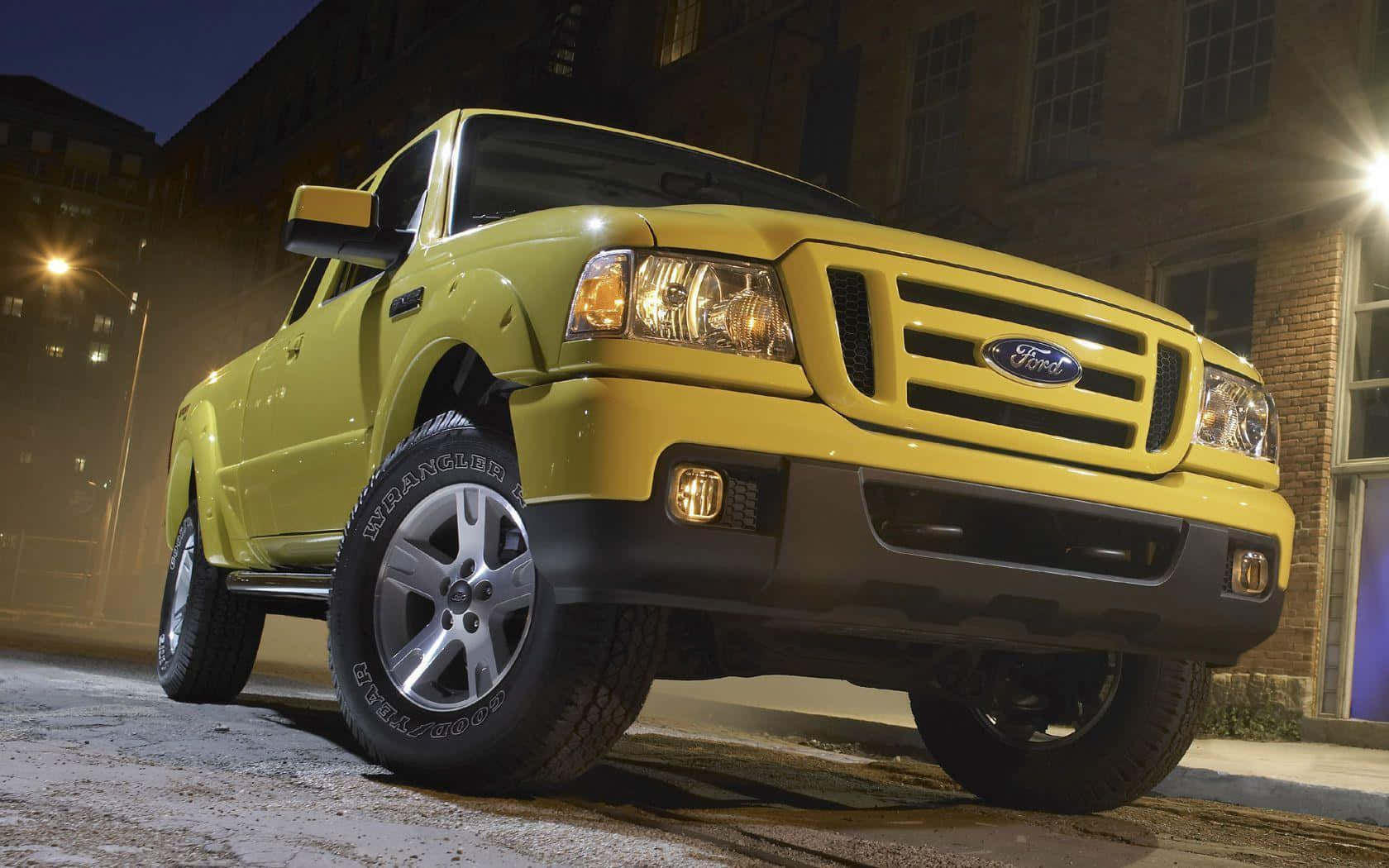 Ford Ranger 桌布 壁紙 — 一輛鮮黃色的福特皮卡在夜間停車，車頭燈亮著，光線投射在磚砌建築附近潮濕的路面上。