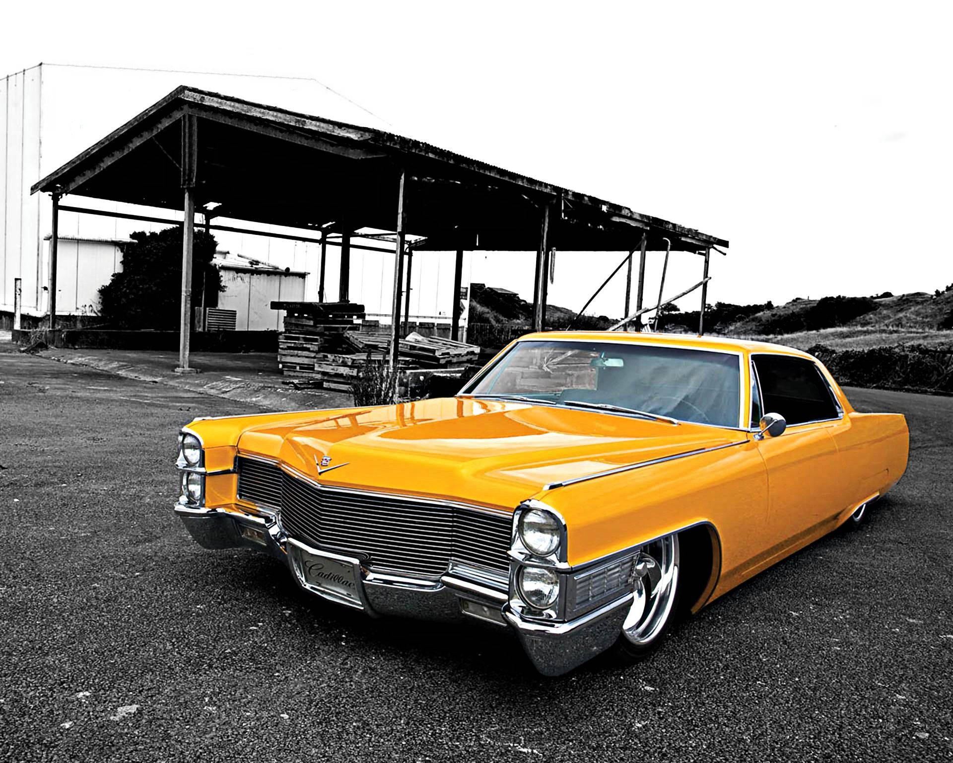 Cadillac 桌布 壁紙 — 一輛鮮黃色的經典凱迪拉克低底盤車在日光下閃耀，停在一個破舊的金屬棚下，位於粗糙的工業區。