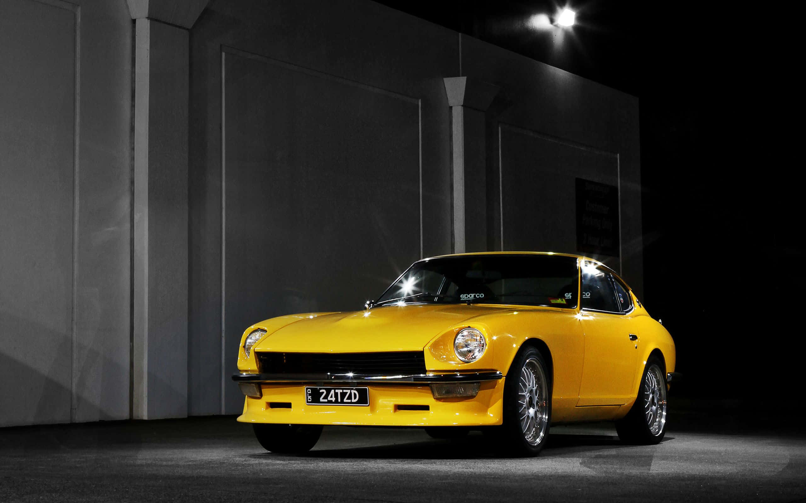 Datsun 桌布 壁紙 — 一輛鮮黃色的經典跑車在夜間單一聚光燈下，在光禿禿的深色混凝土牆前閃閃發光。