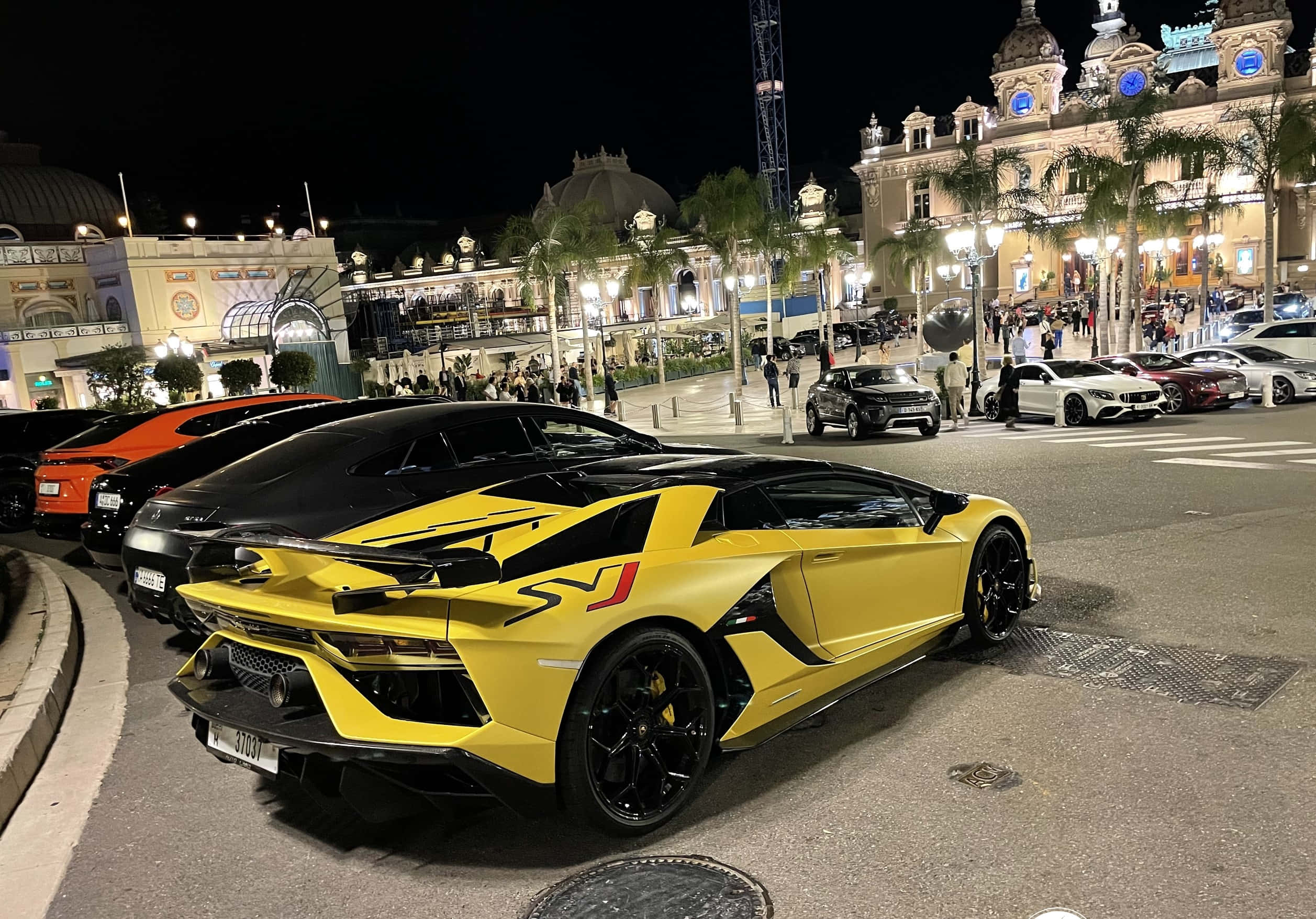Lamborghini Aventador 桌布 壁紙 — 一輛鮮黃色的藍寶堅尼 Aventador SVJ 在夜晚停泊在繁華的豪華度假村，周圍有燈光璀璨的建築物和棕櫚樹。