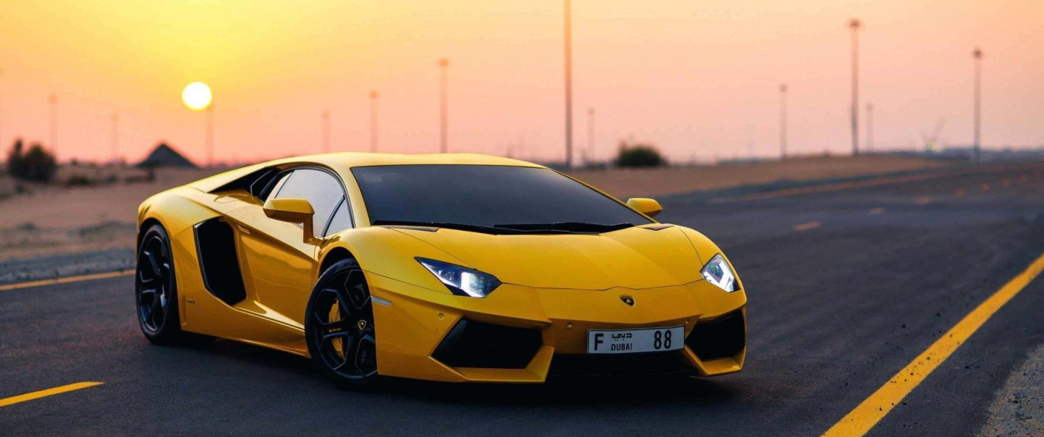 3440x1440p Lamborghini 桌布 壁紙 — 一輛鮮黃色的藍寶堅尼 Aventador 在日落時分沿沙漠公路飛馳，車頭燈閃爍，天空呈現溫暖的橙色。
