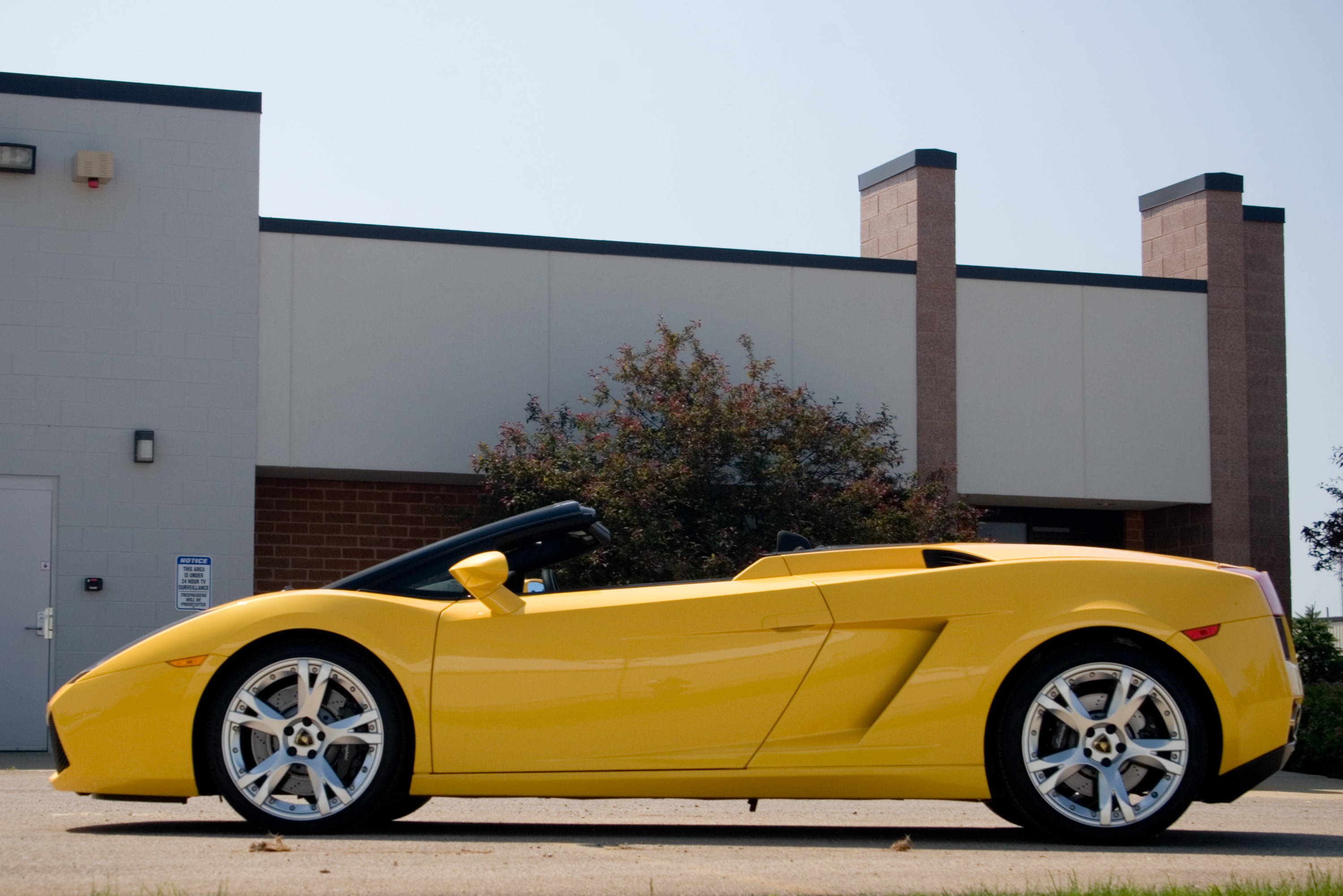 4K 藍寶堅尼 桌布 壁紙 — 一輛鮮黃色的藍寶堅尼 Gallardo Spyder 停泊在現代建築物前的柏油路上，天空晴朗。
