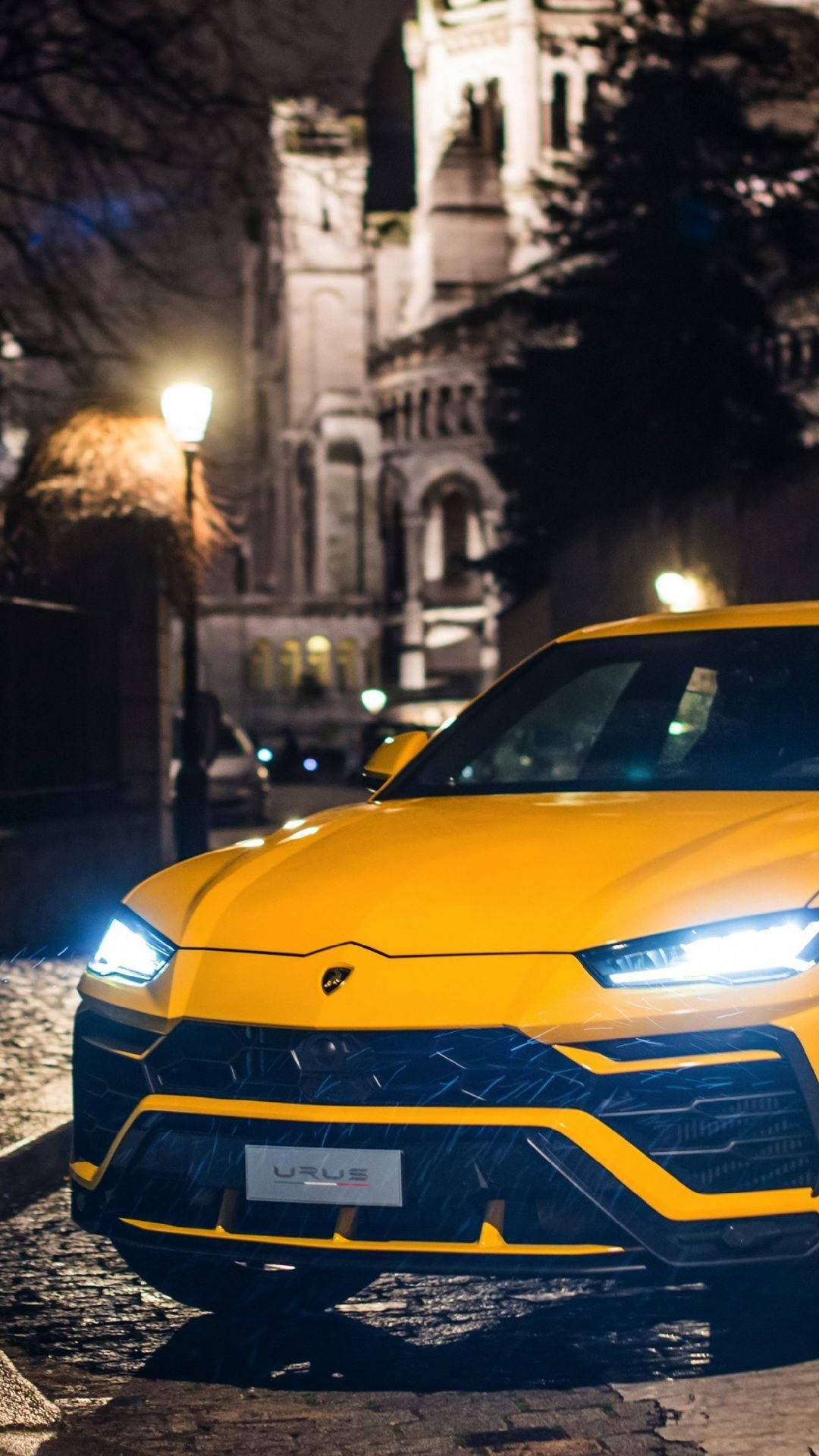 lambo 桌布 壁紙 — 一輛鮮黃色的藍寶堅尼 Urus 在夜間濕滑的鵝卵石街道上，在街燈下閃閃發光，背景是一座歷史建築。