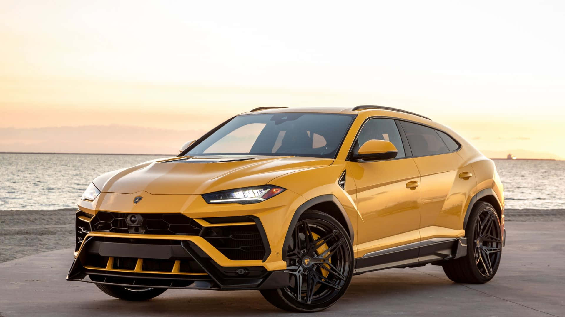 Lamborghini Urus 桌布 壁紙 — 一輛鮮黃色的藍寶堅尼 Urus SUV 停泊在日落時分的混凝土碼頭上，背景是海浪。