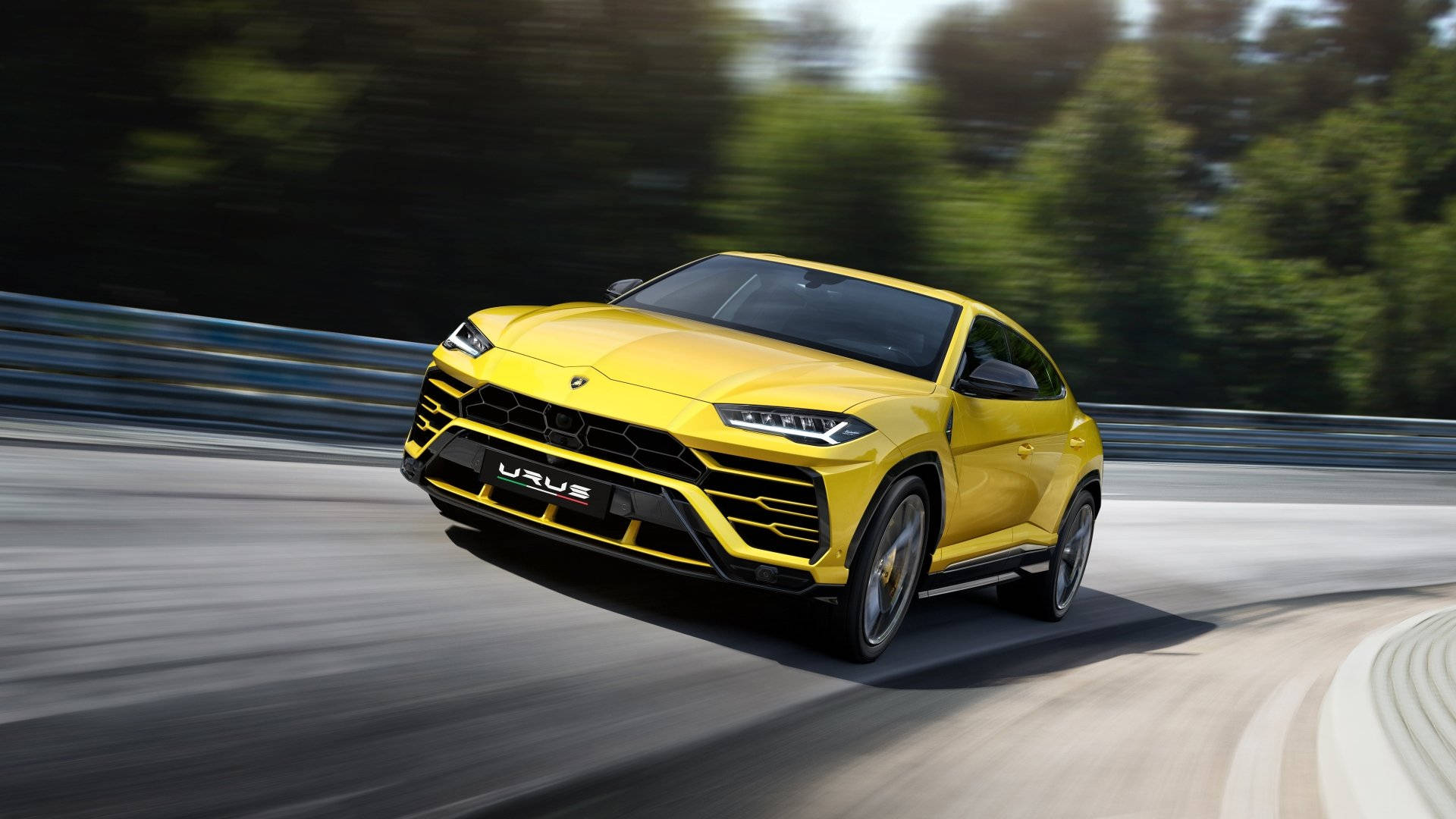 lambo 桌布 壁紙 — 一輛鮮黃色的藍寶堅尼 Urus 沿著彎曲的道路疾馳，背景是模糊的綠色森林。
