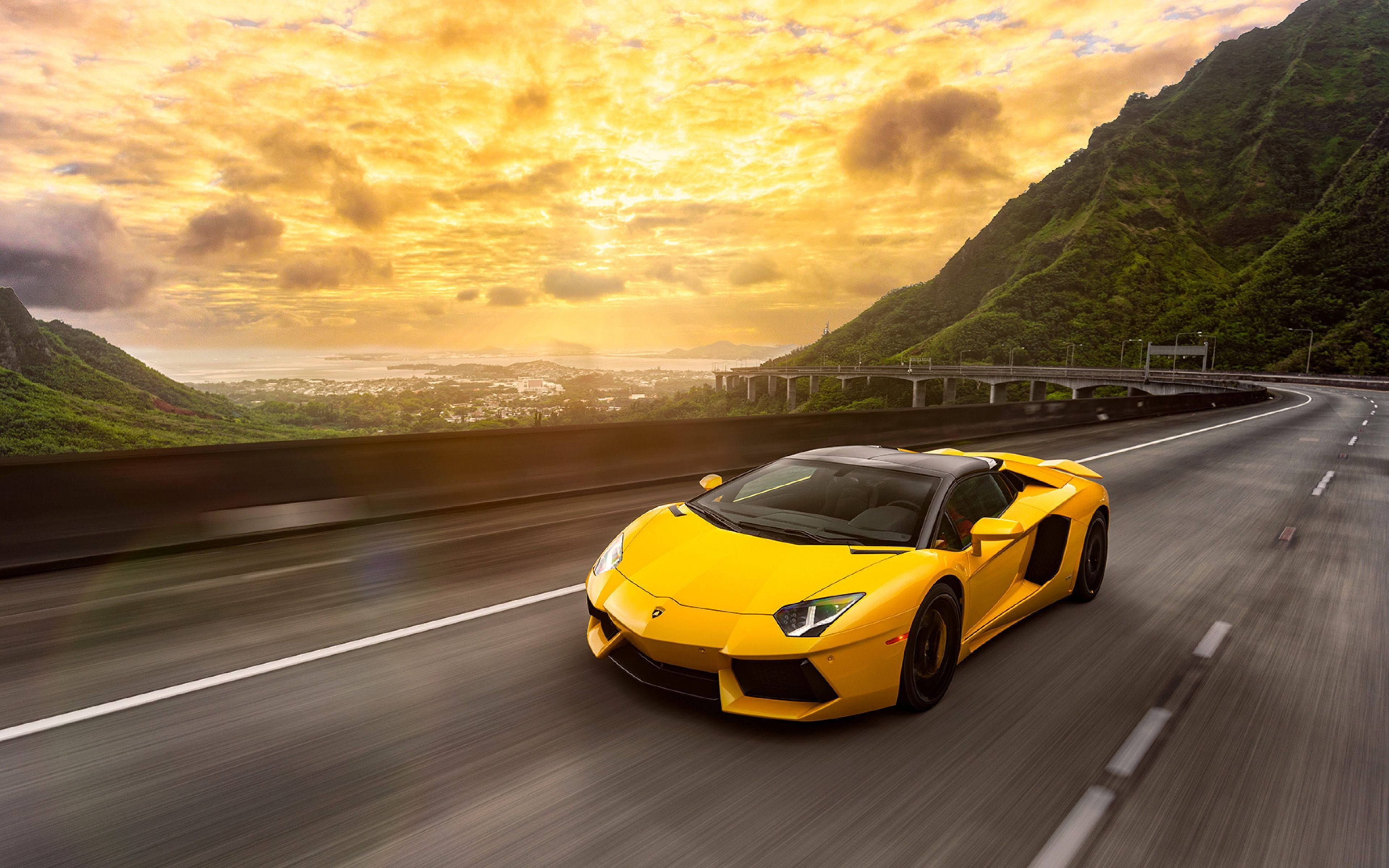 汽車 4K 桌布 壁紙 — 一輛鮮黃色的藍寶堅尼在金色夕陽天空下沿著蜿蜒的山路飛馳。