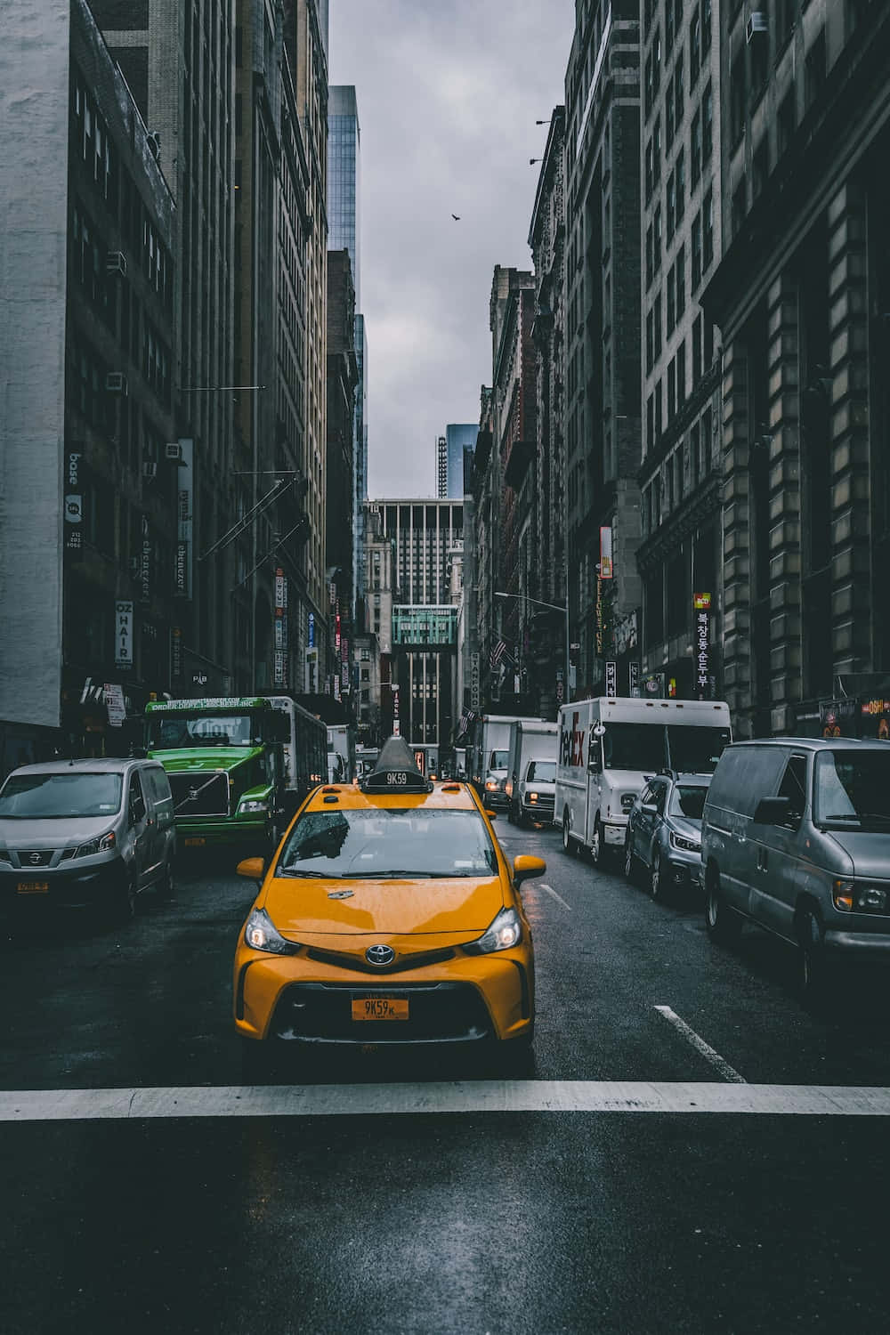 Yellow Cab 桌布 壁紙 — 一輛鮮黃色的計程車在陰天之下，沿著兩旁高聳摩天大樓的紐約市濕滑街道行駛。