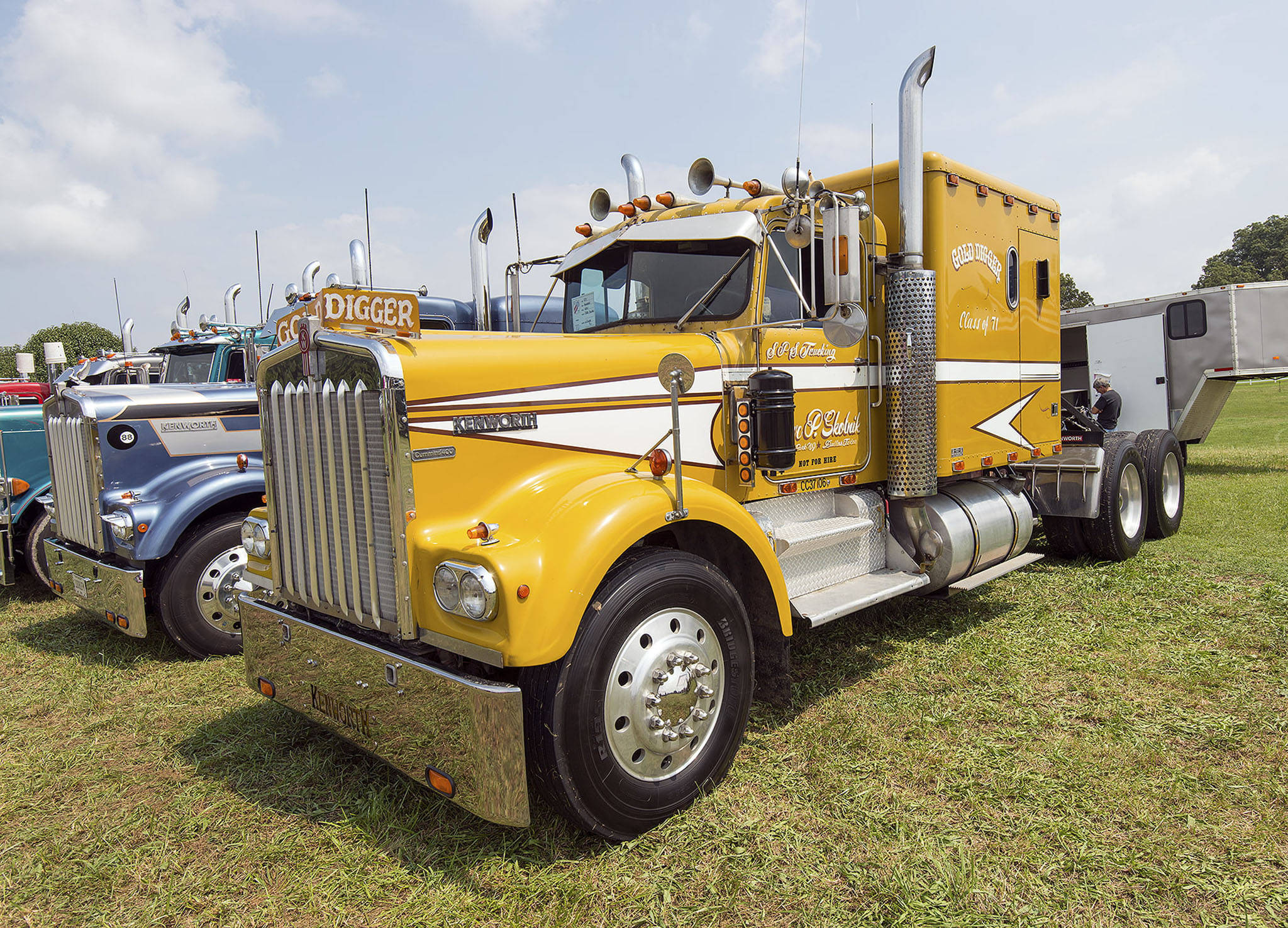 Kenworth W900 桌布 壁紙 — 一輛鮮黃色經典半掛卡車，帶有鍍鉻細節和定制貼花，停在卡車展的草地上，天空部分多雲。