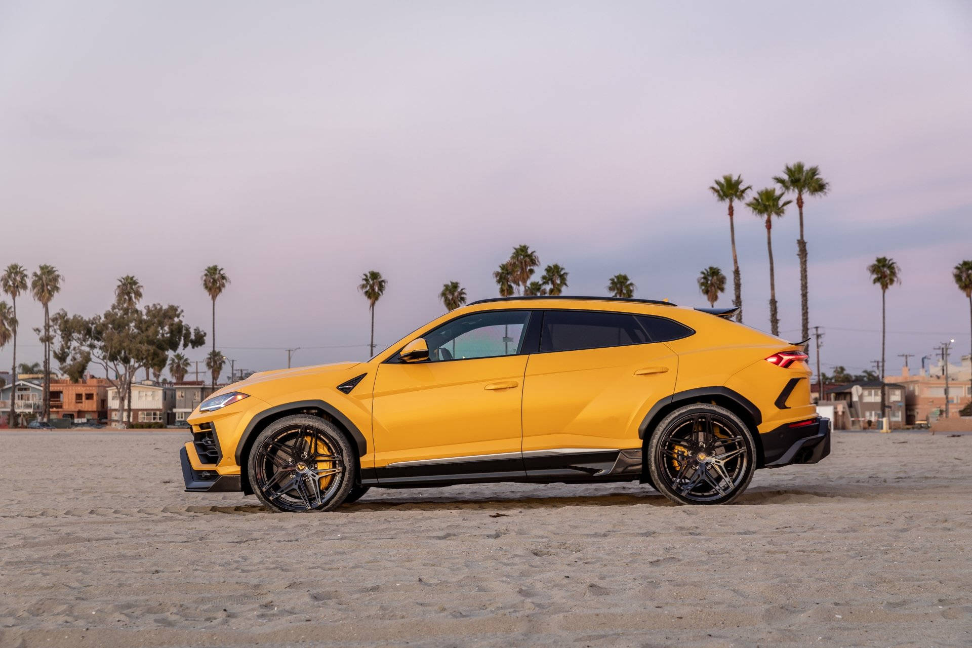 lambo 桌布 壁紙 — 一輛鮮黃色藍寶堅尼 Urus SUV 停放在沙灘上，背景有棕櫚樹和柔和的暮色天空。
