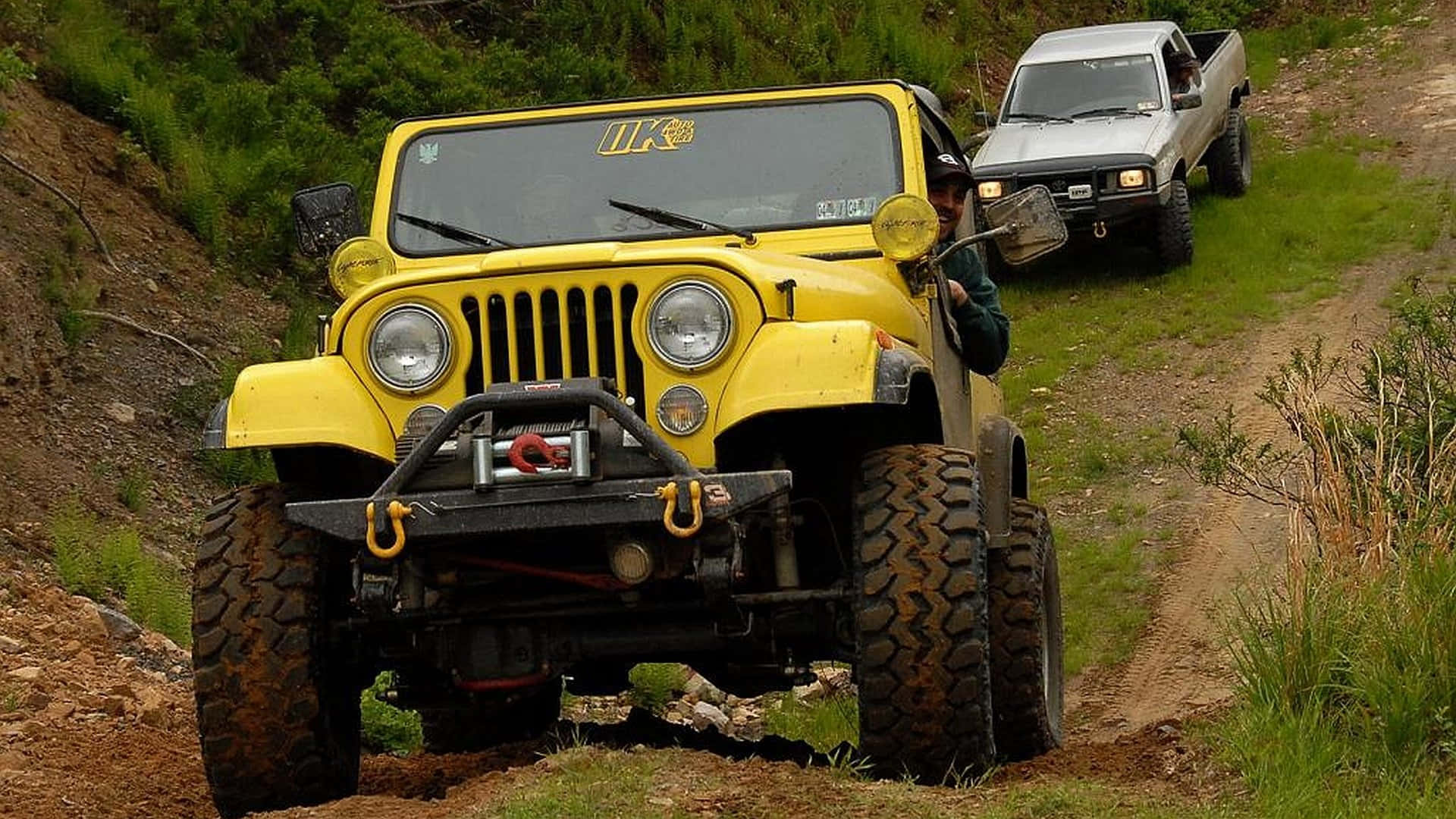 Jeep Cj 桌布 壁紙 — 一輛黃色Jeep Wrangler配備大型輪胎，正在攀爬泥濘的山坡小徑，後面跟著一輛銀色皮卡。