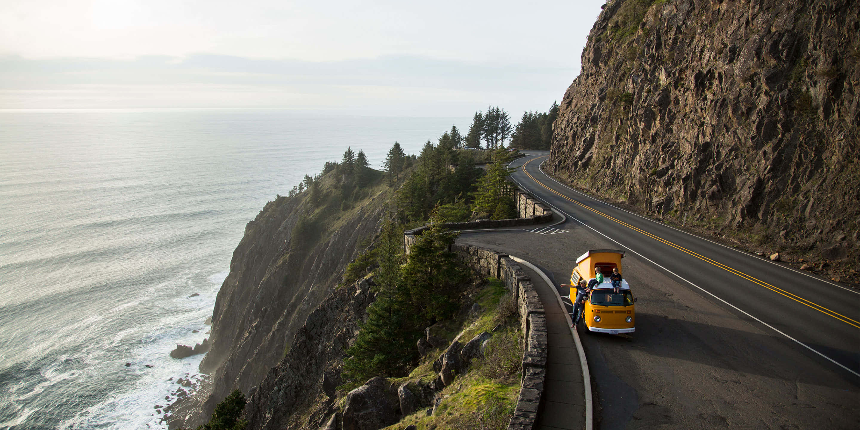 Oregon Coast 桌布 壁紙 — 一輛黃色復古廂型車沿著蜿蜒的海岸道路行駛，一側是懸崖和海洋，另一側是森林覆蓋的山丘。