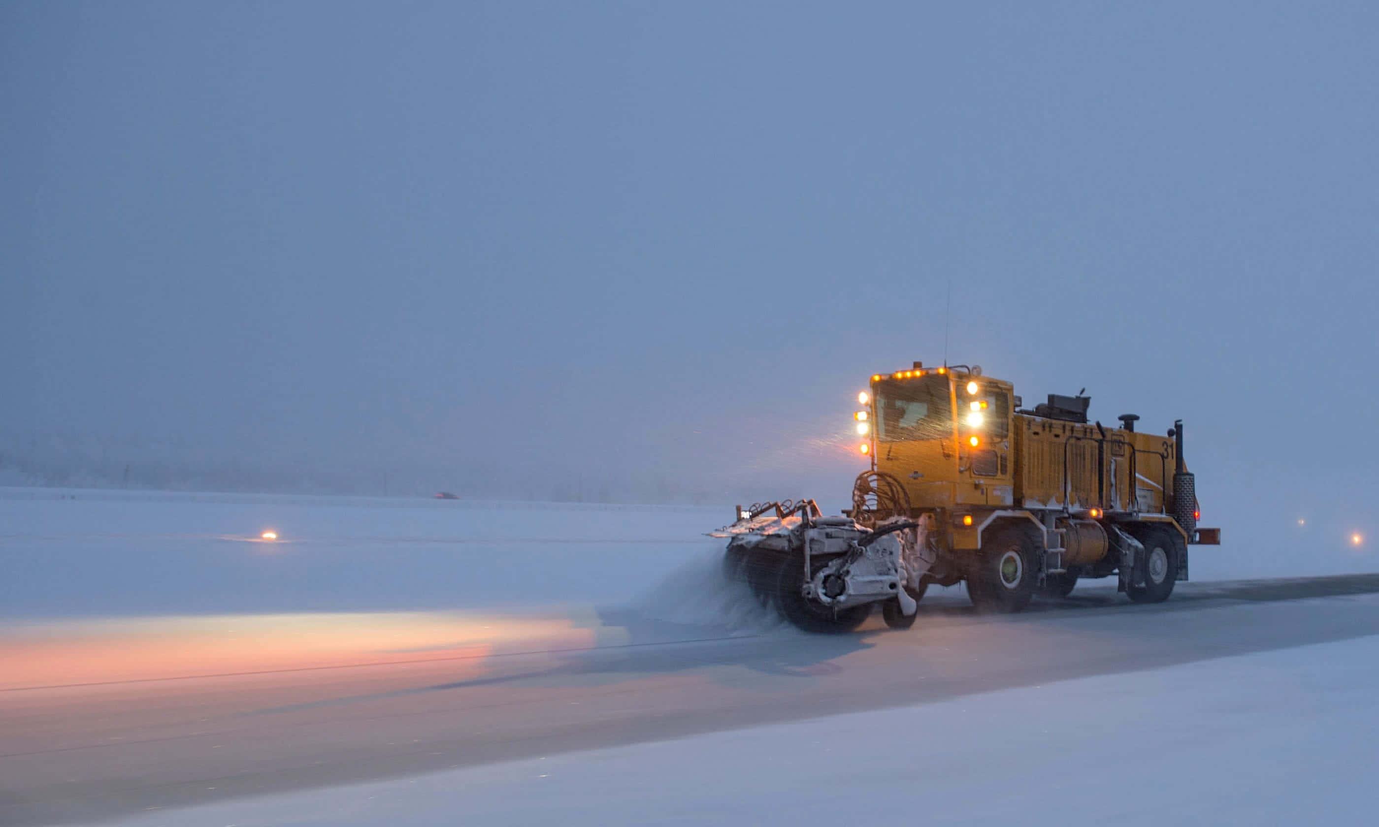 Snowplow 桌布 壁紙 — 一輛黃色掃雪車在黃昏時清理積雪的道路，車頭燈照亮飄落的雪花。