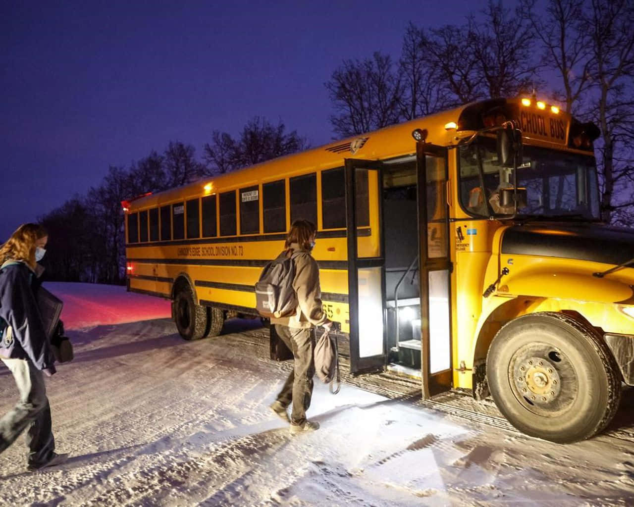 Bus 桌布 壁紙 — 一輛黃色校車停放在雪暮中，學生穿著冬季裝備在其明亮的車內燈光下上車。