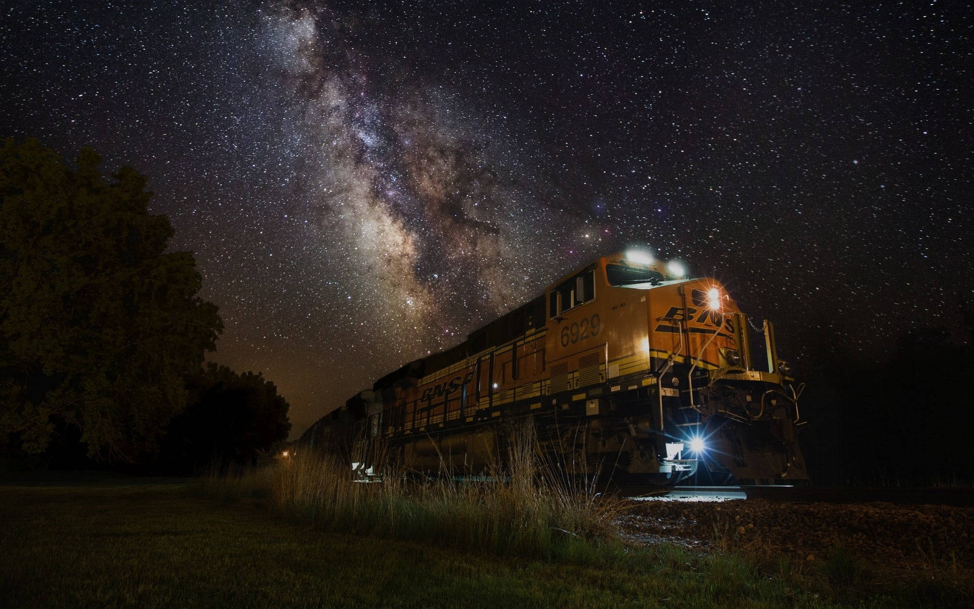 夜晚銀河星下的黃色火車，伴隨著草地和樹木。