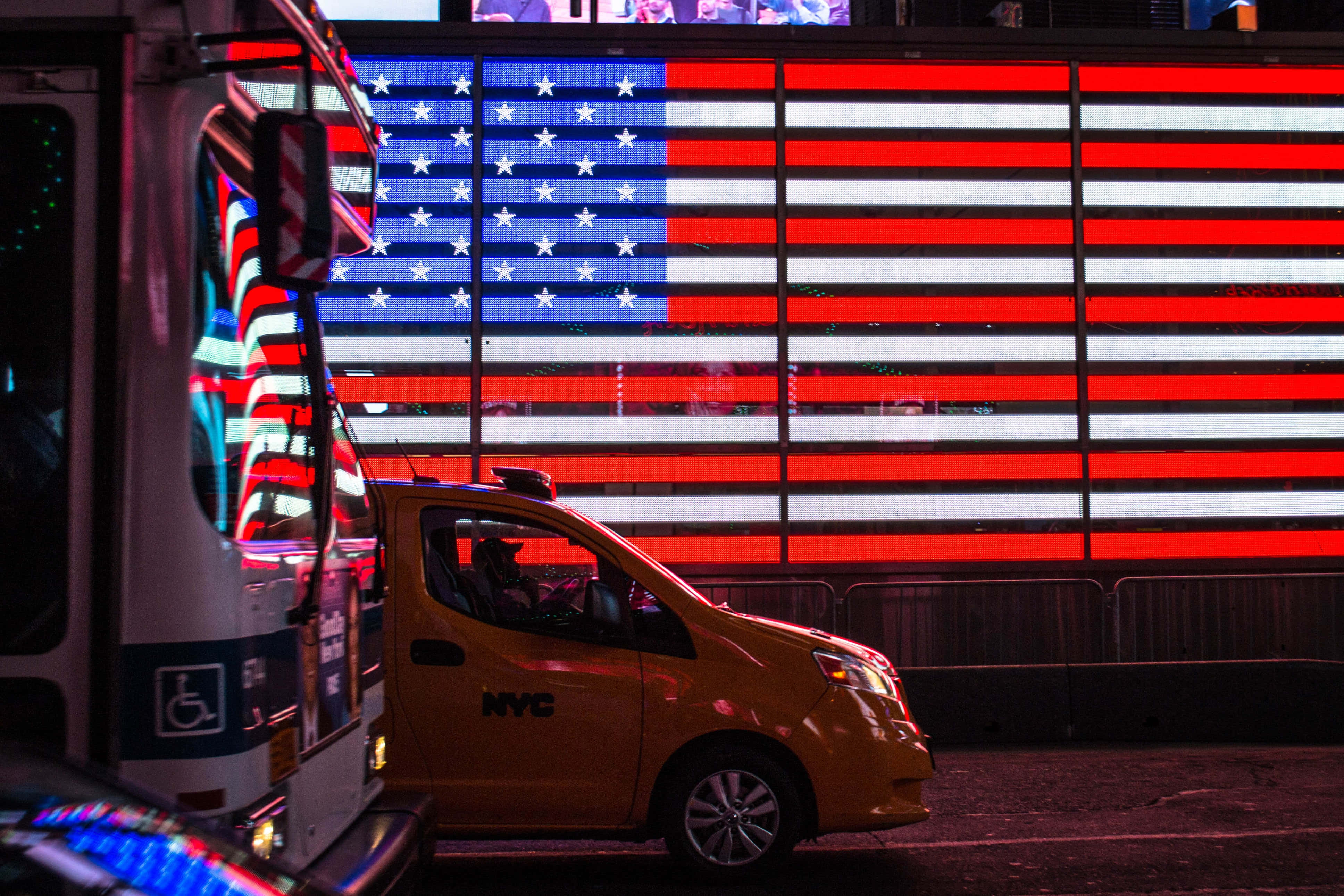 US Flag 桌布 壁紙 — 一輛黃色的紐約市計程車和救護車在夜間停在一棟城市建築上展示的巨大發光美國國旗前。