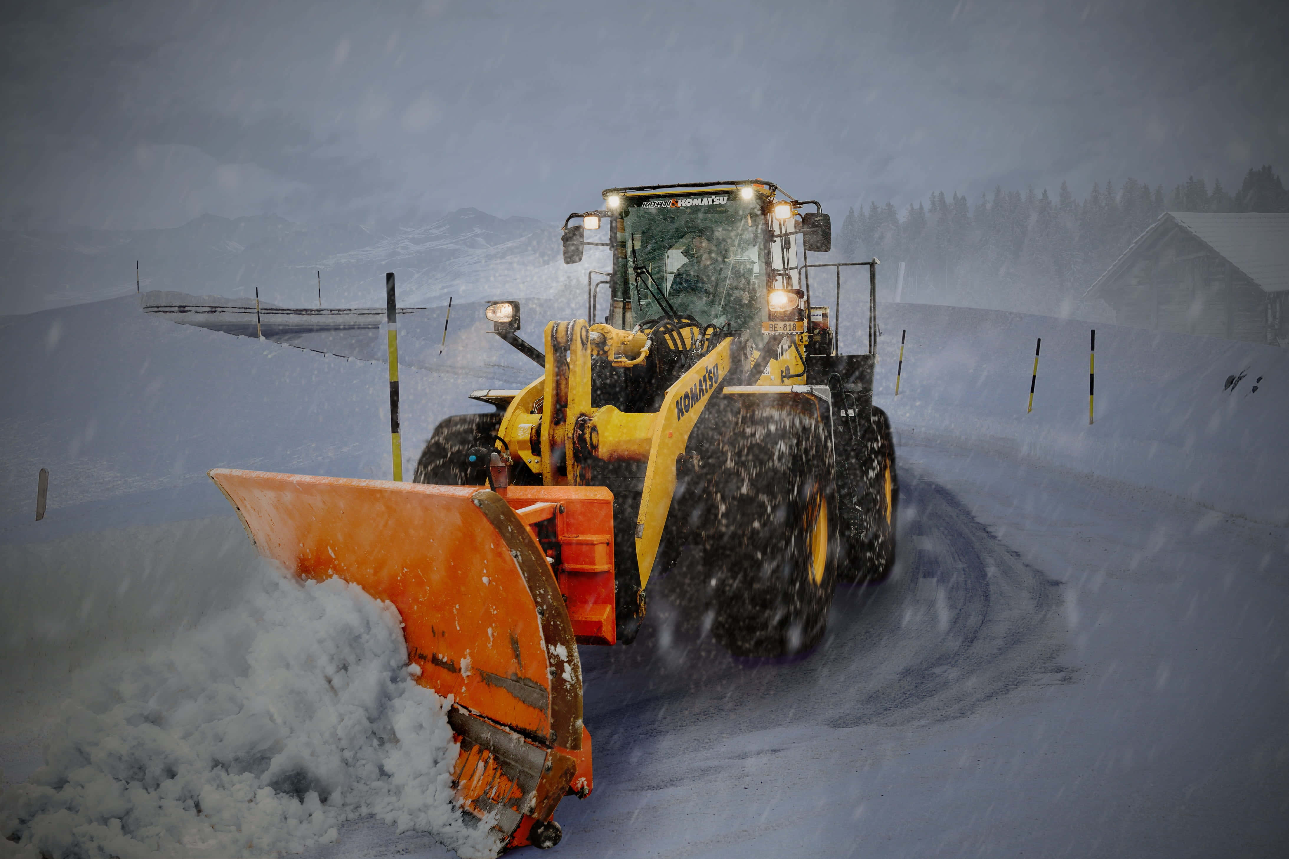 Snowplow 桌布 壁紙 — 一輛黃色鏟雪車在暴雪中清理積雪道路，背景有山脈和小屋。