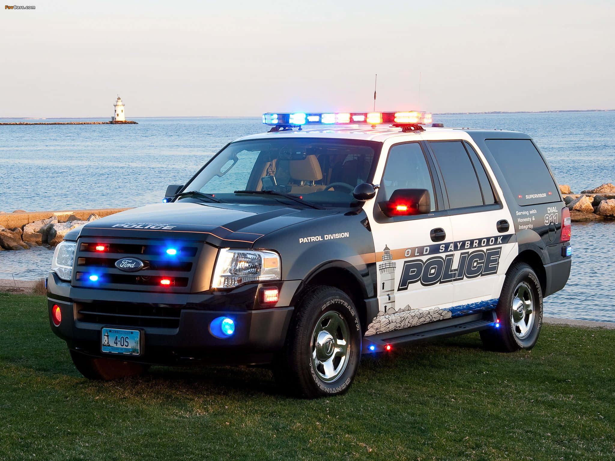 Ford Expedition 桌布 壁紙 — 一輛黑白相間的 Old Saybrook 警用 Ford Explorer SUV，閃爍著藍色和紅色燈光，停在靠近水邊的草地上，遠處有一座燈塔。