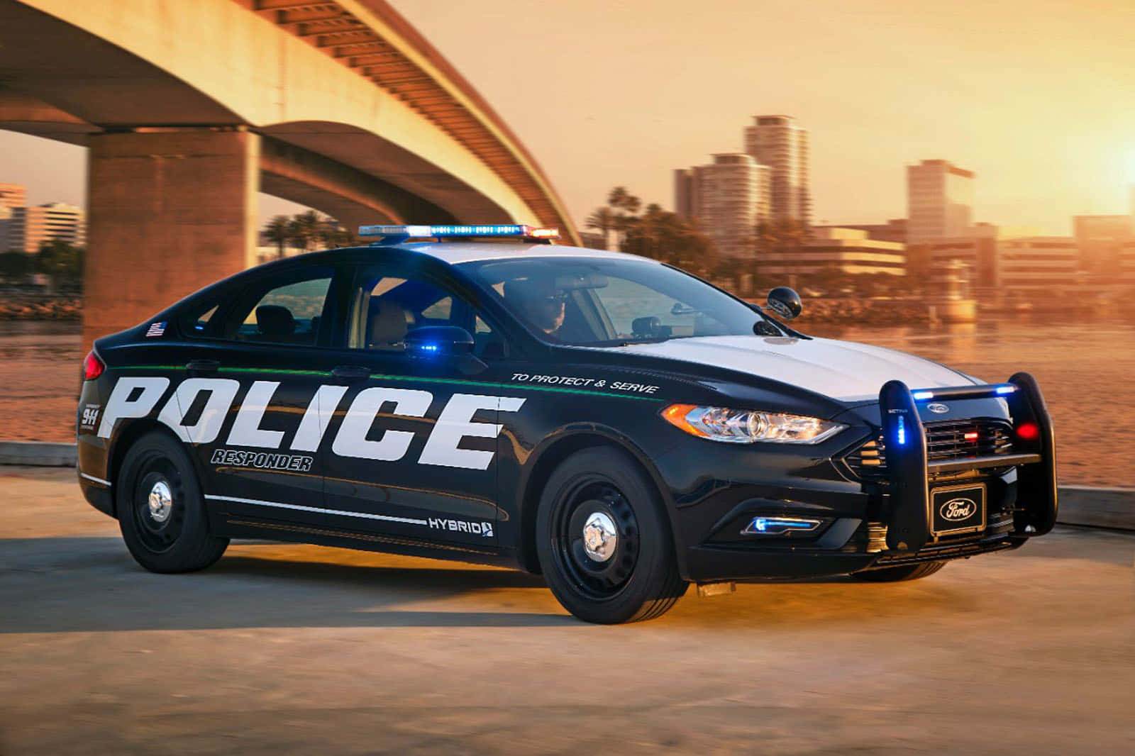 Police Car 桌布 壁紙 — 一輛黑白相間的福特警用混合動力車，閃爍著藍色警燈，在黃金時刻行駛於橋下，背景是城市天際線。