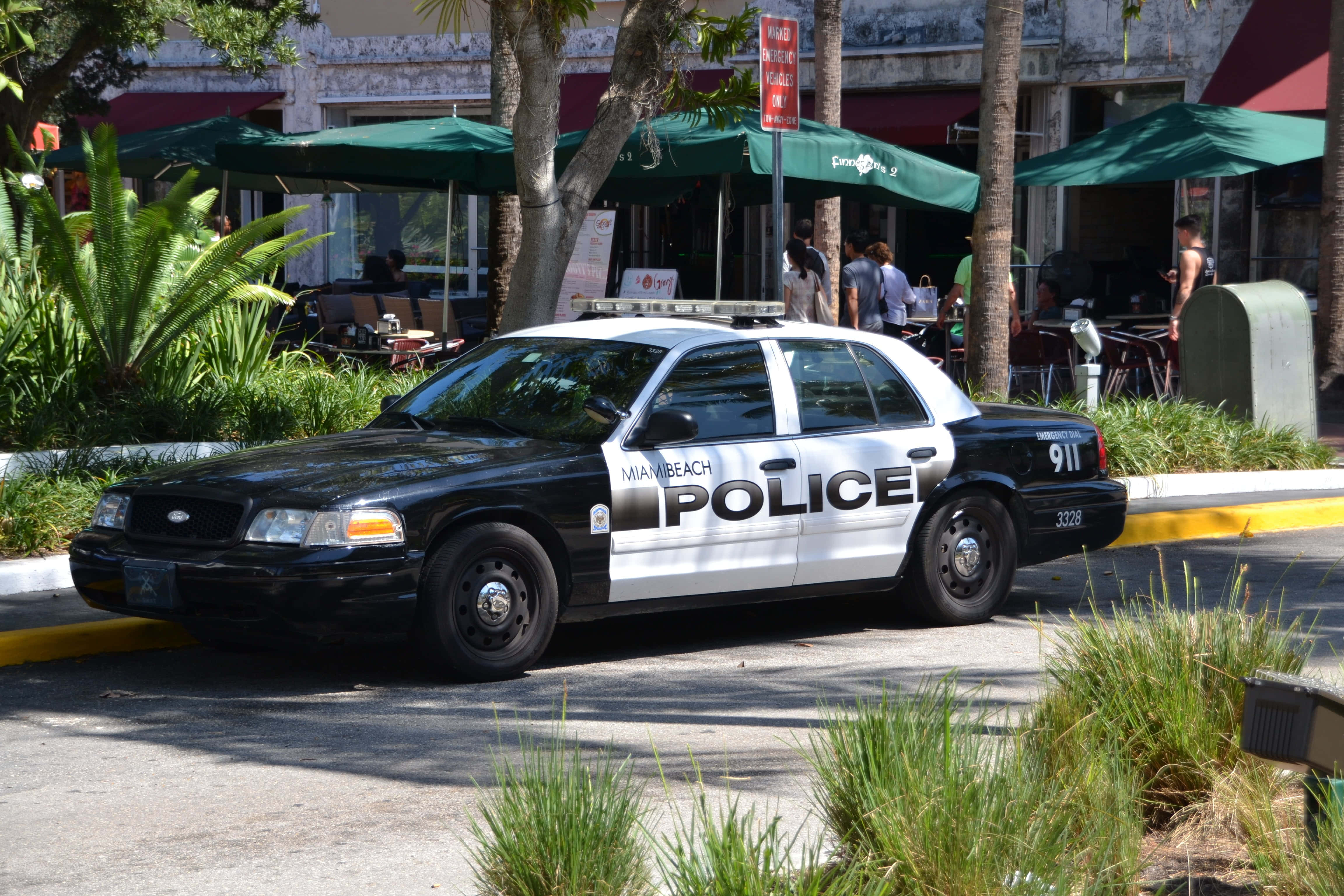 Ford Crown Victoria 桌布 壁紙 — 一輛黑白相間的邁阿密海灘警車停泊在陽光明媚的街道上，街道兩旁種滿棕櫚樹和戶外咖啡館的遮陽傘。