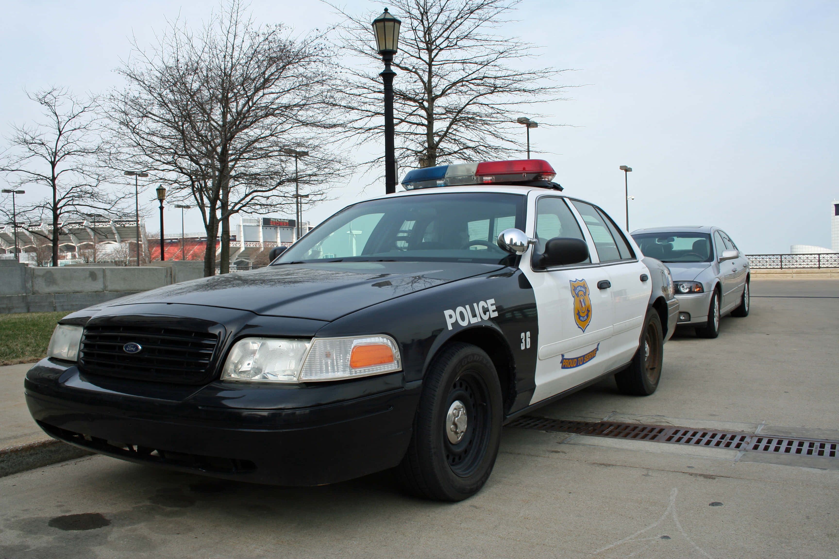 Police Car 桌布 壁紙 — 一輛黑白警車，帶有紅色和藍色燈光，停在鋪砌的停車場，天空多雲。