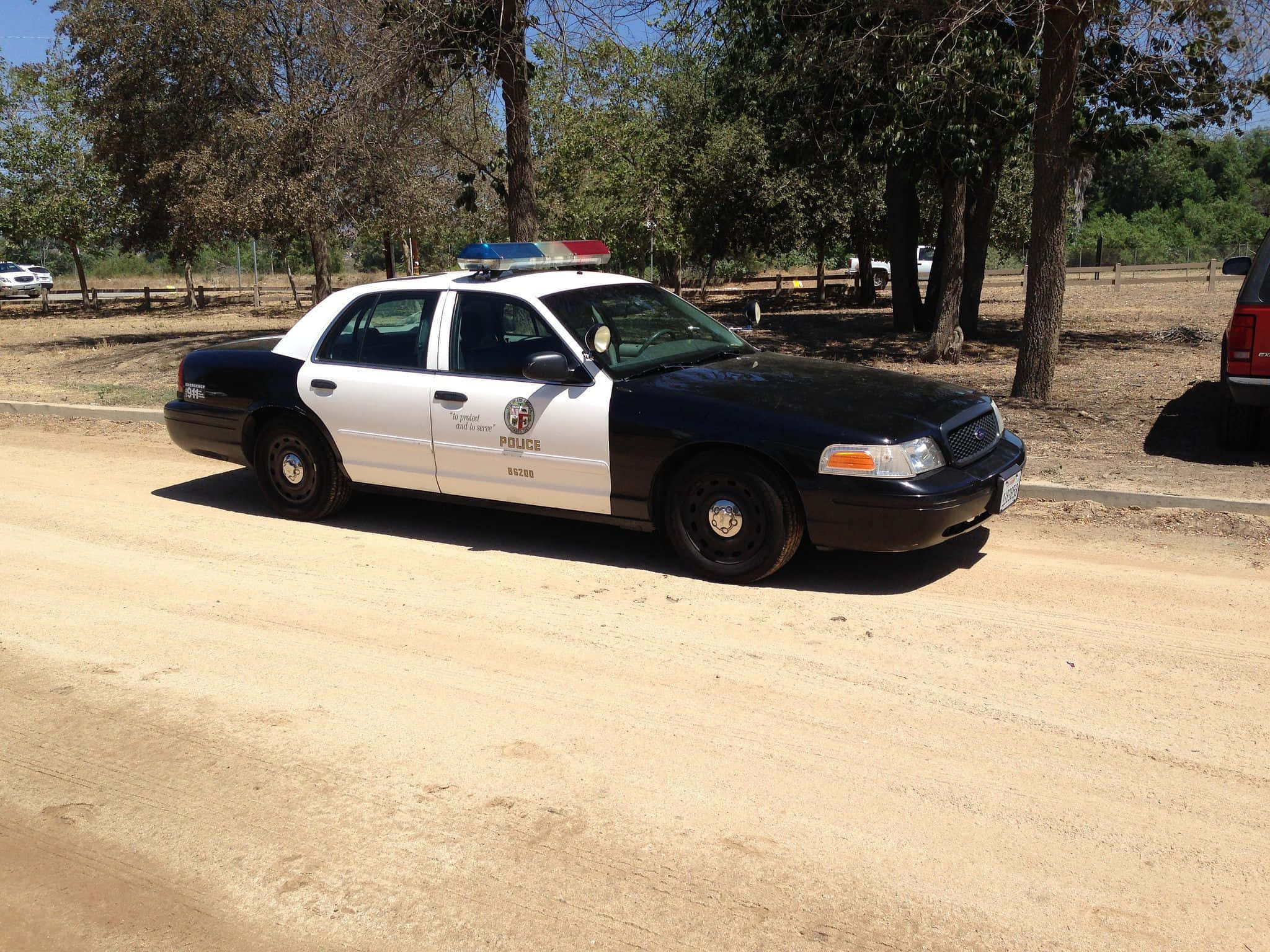 Ford Crown Victoria 桌布 壁紙 — 一輛黑白警車，帶有藍色和紅色燈光，停在塵土飛揚的鄉村道路上，周圍樹木環繞，陽光明媚。