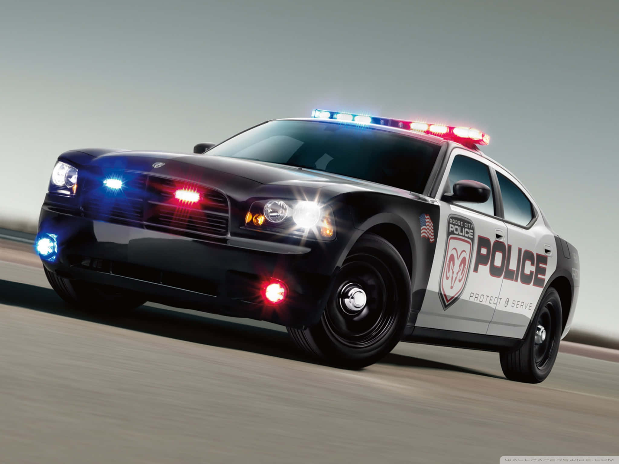 Law Enforcement 桌布 壁紙 — 一輛黑白警車在模糊的灰色背景前，閃爍著紅藍燈光向前疾馳。