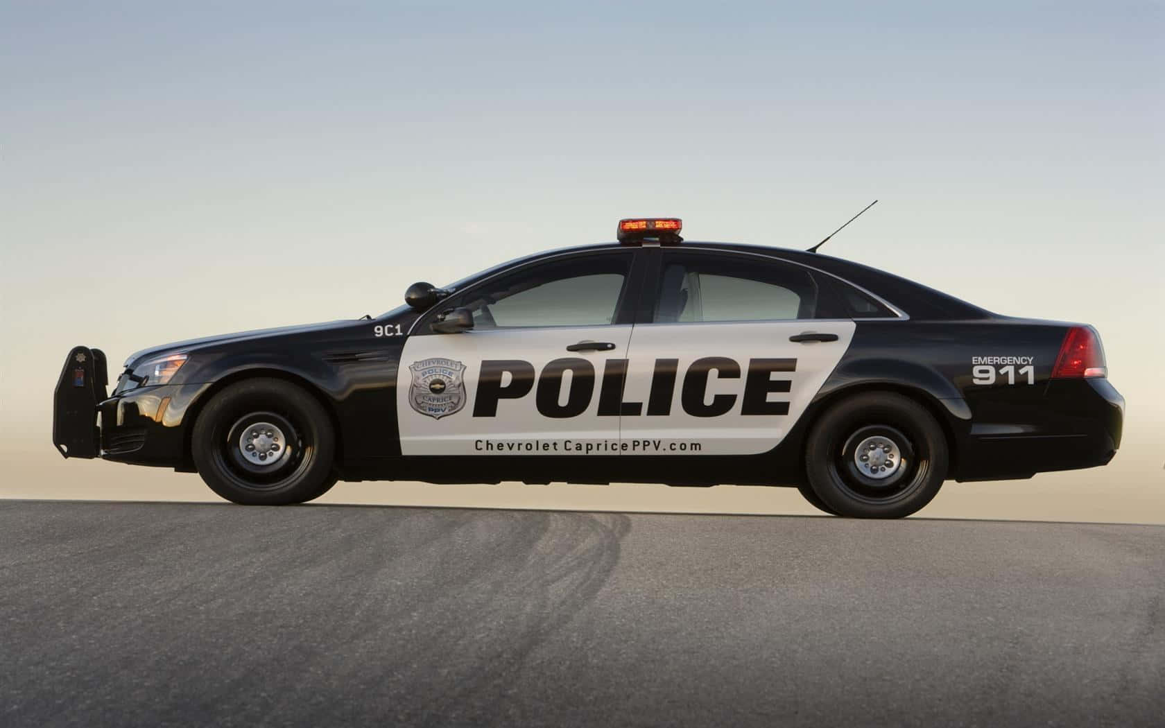 Police Car 桌布 壁紙 — 一輛黑白警車帶有 'POLICE' 和 'EMERGENCY 911' 標記，停在晴朗天空下的瀝青路面上。