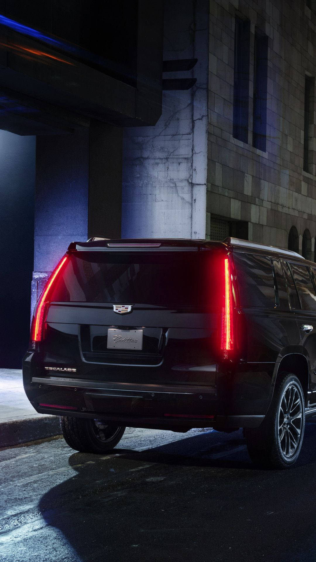 凱迪拉克 iPhone 桌布 壁紙 — 一輛黑色 Cadillac Escalade SUV 在夜間停在一座石建築前的橋下，散發出鮮豔的紅色尾燈光芒。
