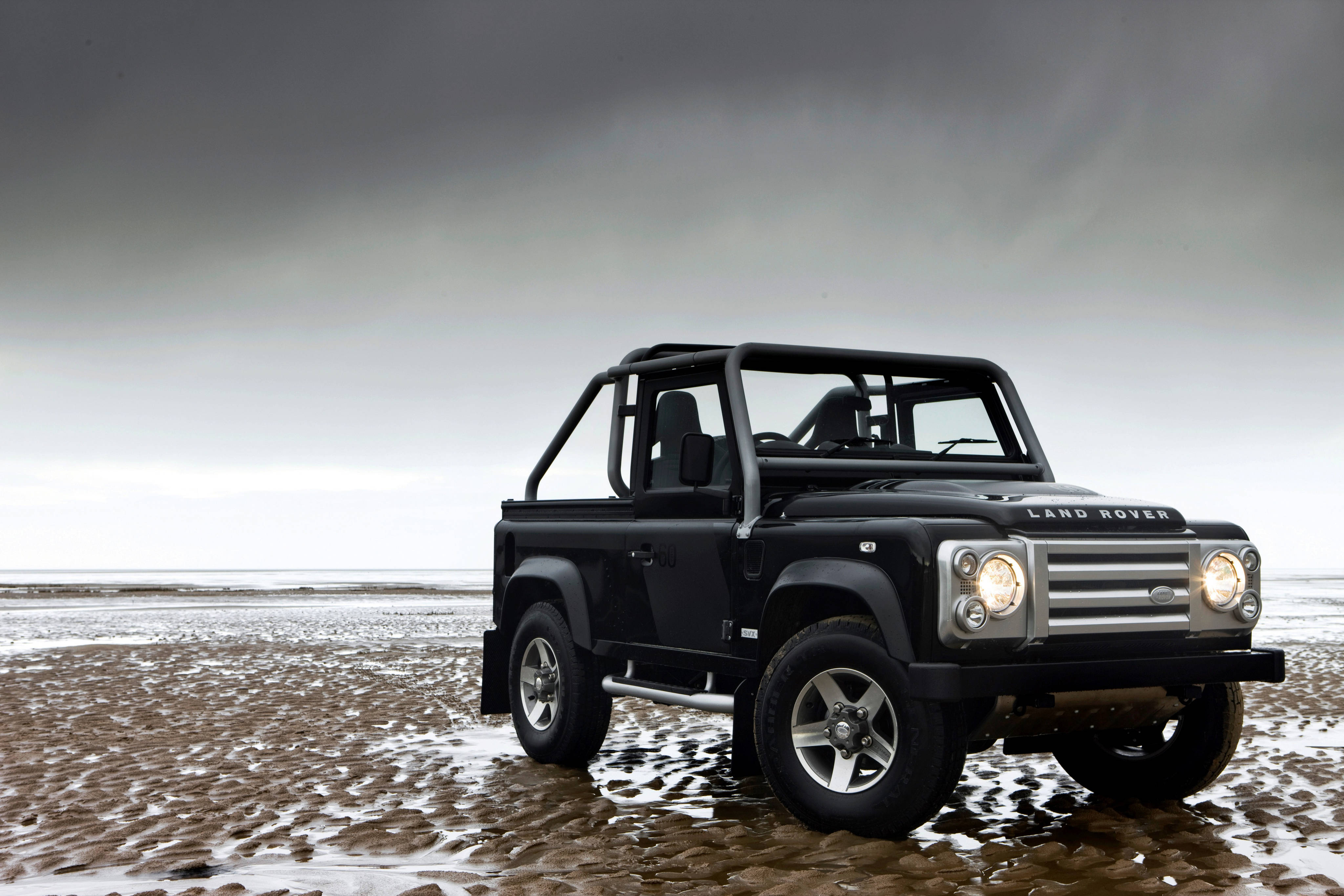 land rover iPhone 桌布 壁紙 — 一輛黑色 Land Rover Defender 在沉重的灰色天空下，堅固地停在濕漉漉的沙灘上。