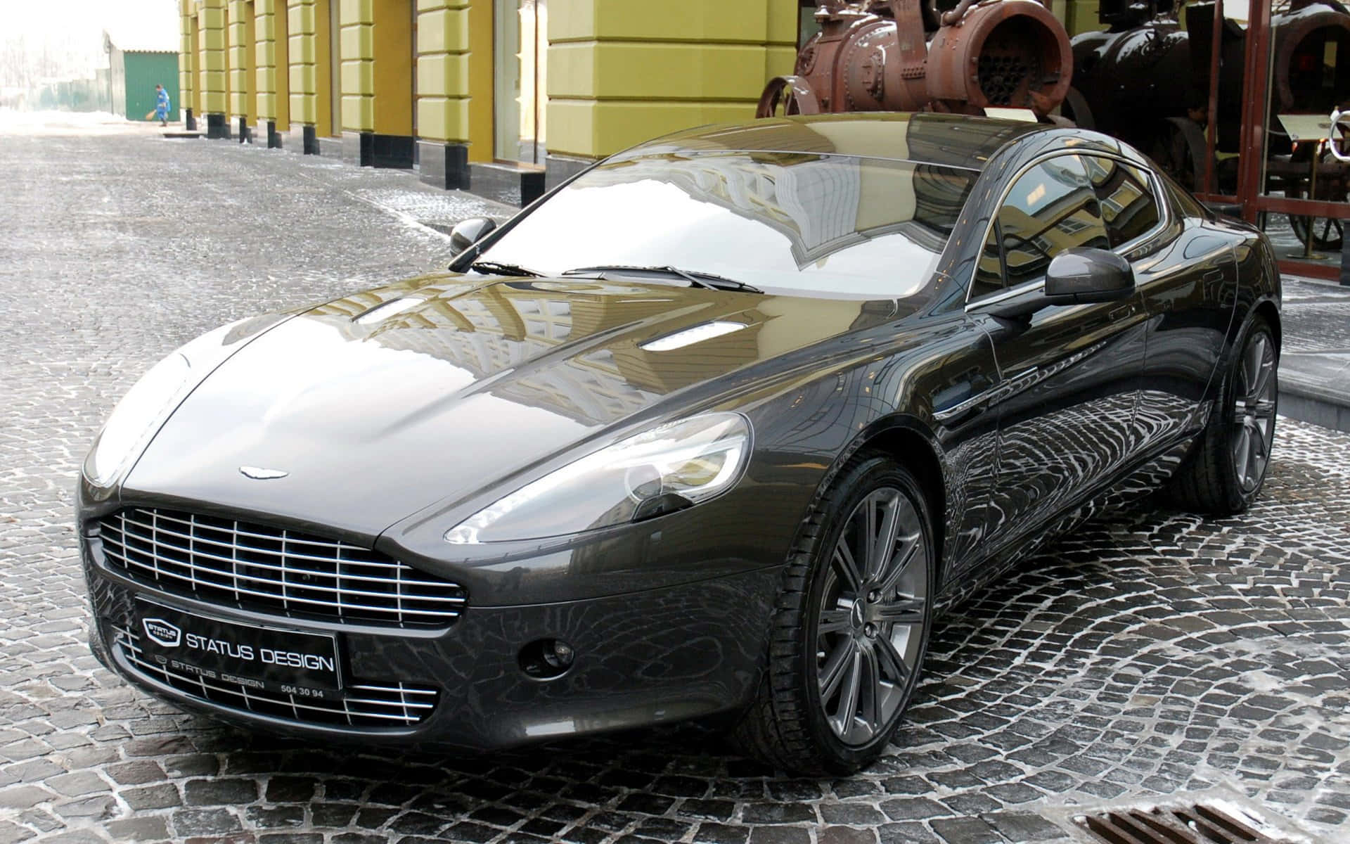 Aston Martin Rapide S 桌布 壁紙 — 一輛黑色Aston Martin跑車在濕滑的鵝卵石街道上閃閃發光，旁邊是一座黃色建築物。
