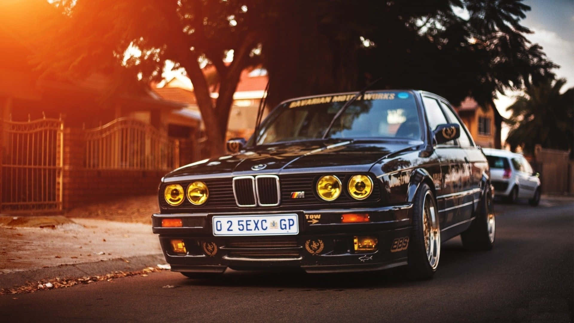 1920x1080 Bmw 桌布 壁紙 — 一輛黑色BMW E30跑車，黃色車頭燈閃爍，停在黃金時段綠樹成蔭的街道上。