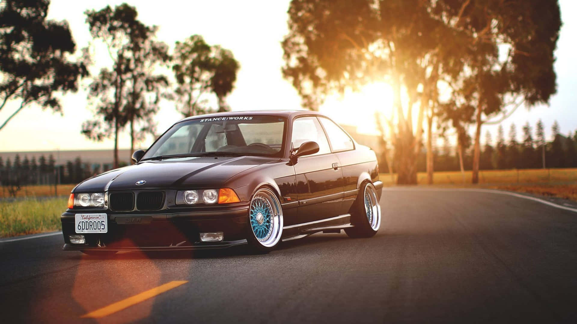 Bmw Cars 桌布 壁紙 — 一輛黑色BMW E36雙門轎跑車搭配藍色輪圈，低趴於日落時分的道路上，樹木剪影在後。