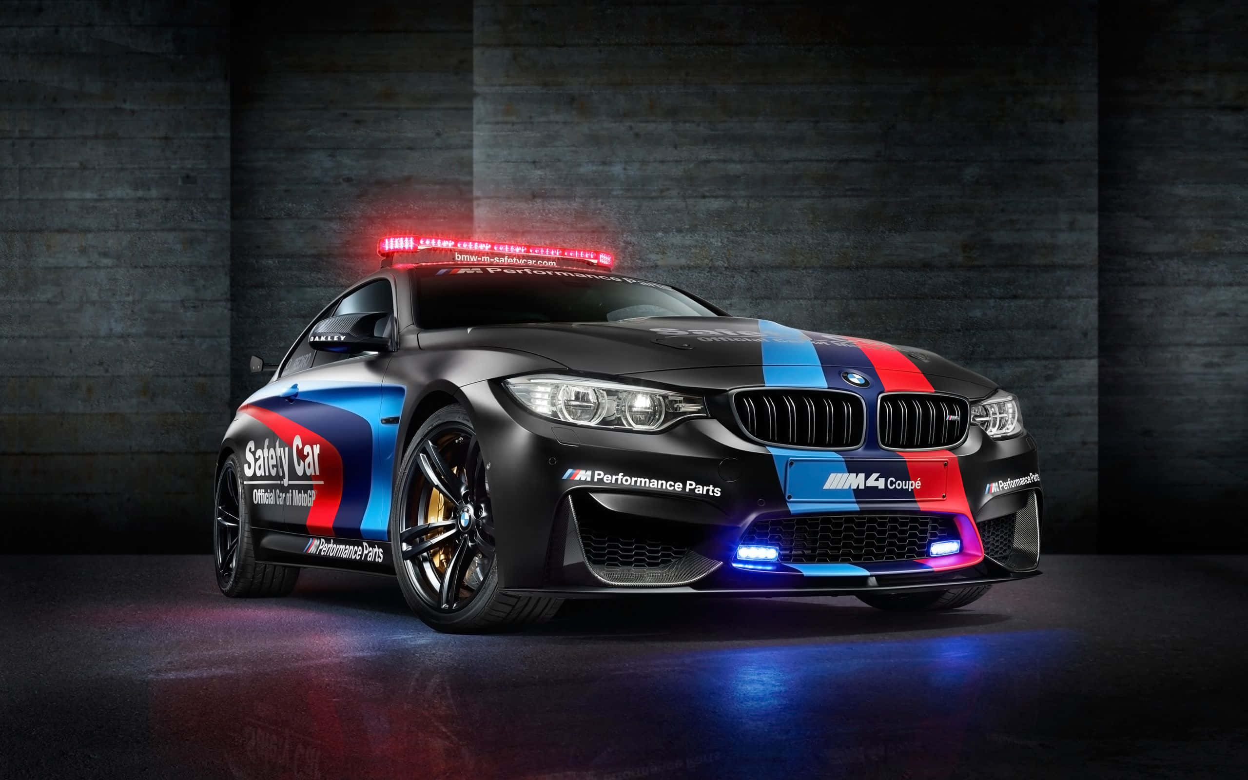 Bmw M Series 桌布 壁紙 — 一輛黑色BMW M4 Coupe安全車，帶有紅、藍、白色賽車條紋和閃燈，停在黑暗的混凝土車庫中。