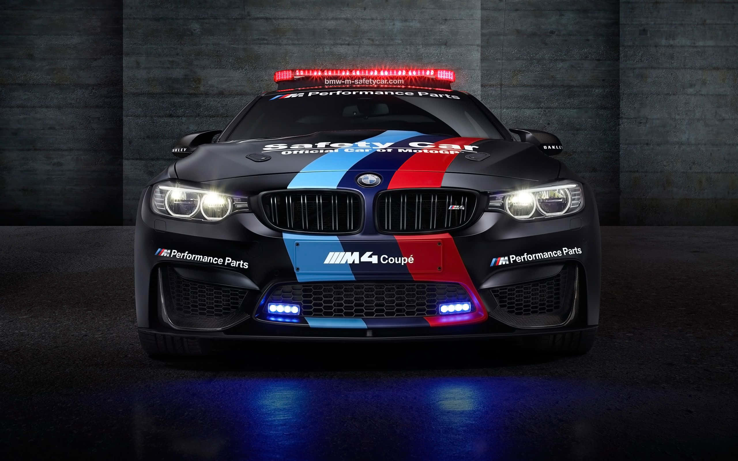 汽車 桌布 壁紙 — 一輛黑色BMW M4 Coupe安全車，帶有紅、藍、白色賽車條紋和閃爍燈光，停在黑暗的混凝土車庫中。
