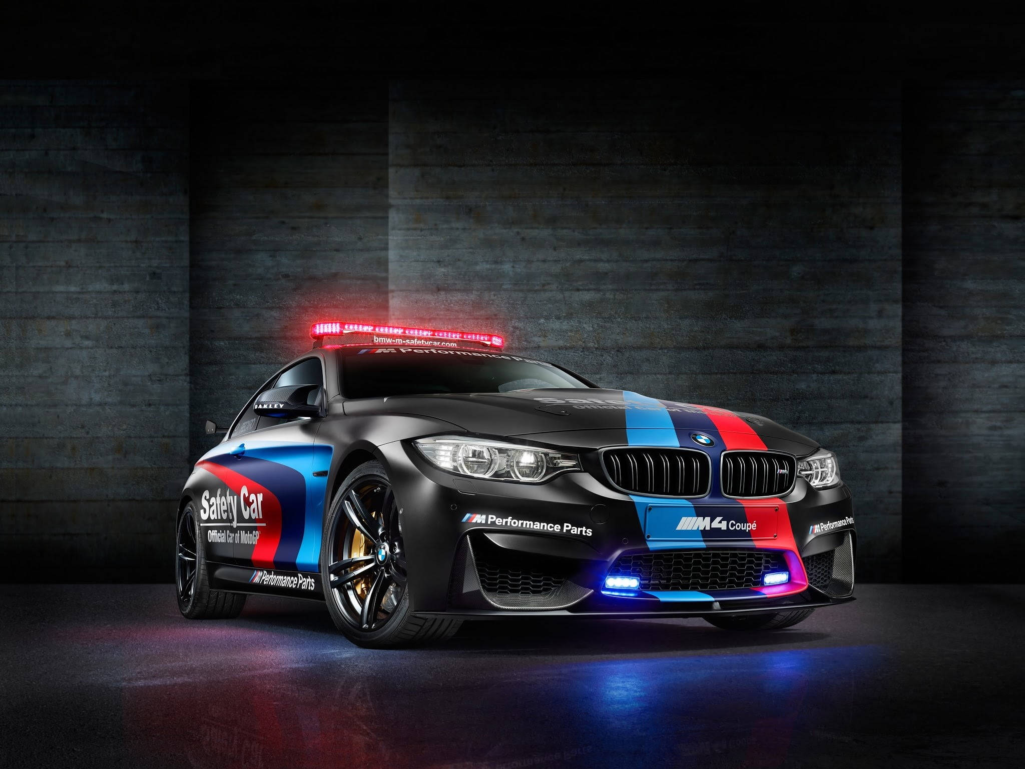 BMW 動圖 桌布 壁紙 — 一輛黑色BMW M4 Coupe安全車，帶有紅、藍、白色賽車條紋，在戲劇性的攝影棚燈光下，映襯著深色混凝土牆壁閃耀。