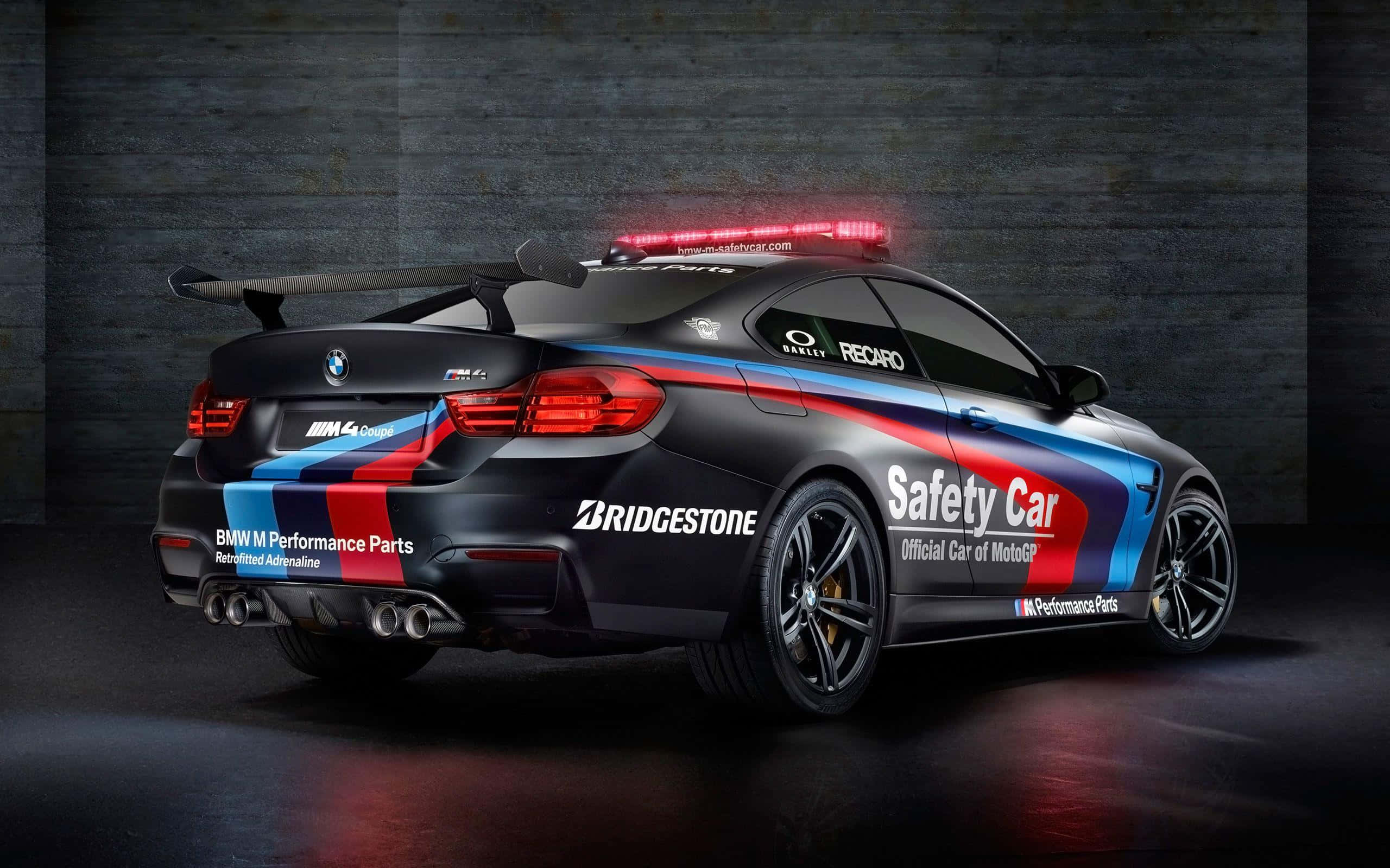 Safety Car 桌布 壁紙 — 一輛黑色BMW M4賽車，帶有紅、藍、白色賽車塗裝和閃爍的紅燈，停在深色混凝土車庫地板上。