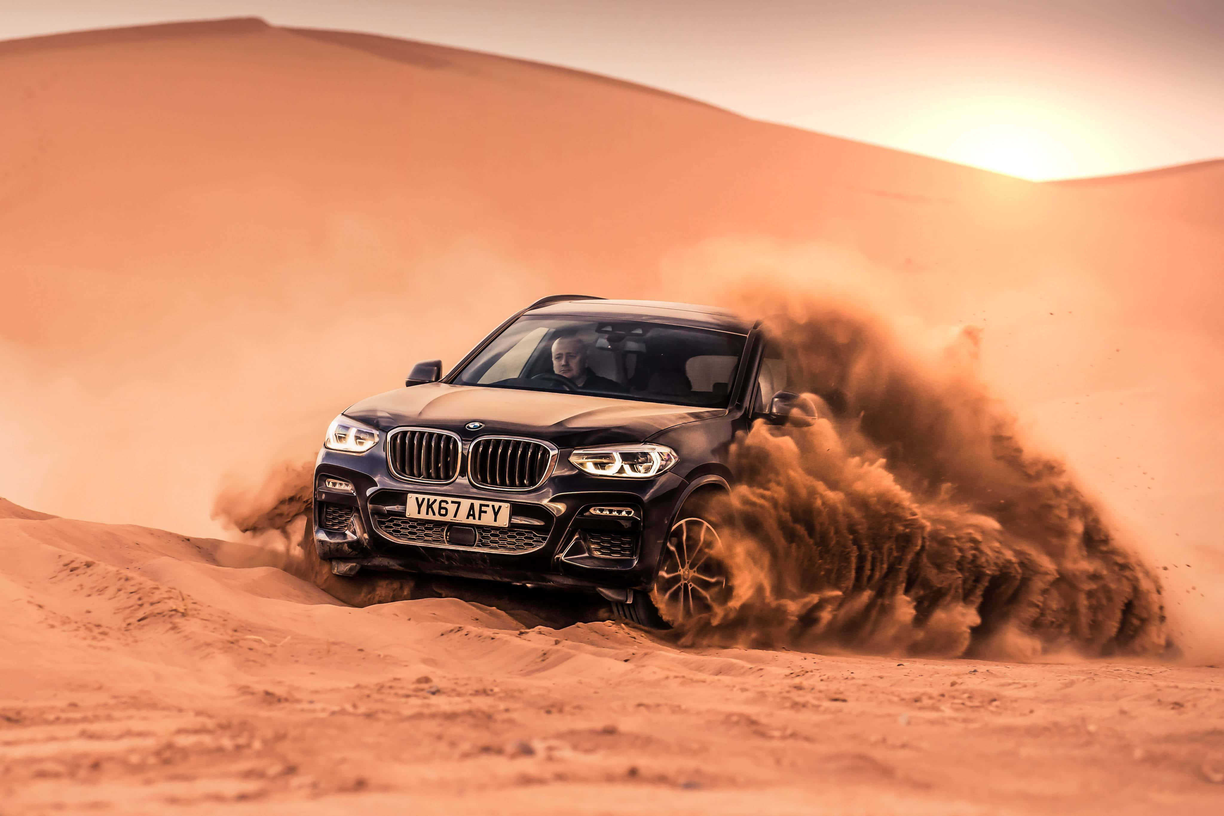 Bmw X3 桌布 壁紙 — 一輛黑色BMW SUV在沙漠沙丘上奔馳，在溫暖的夕陽天空下揚起巨大的橙色沙塵雲。