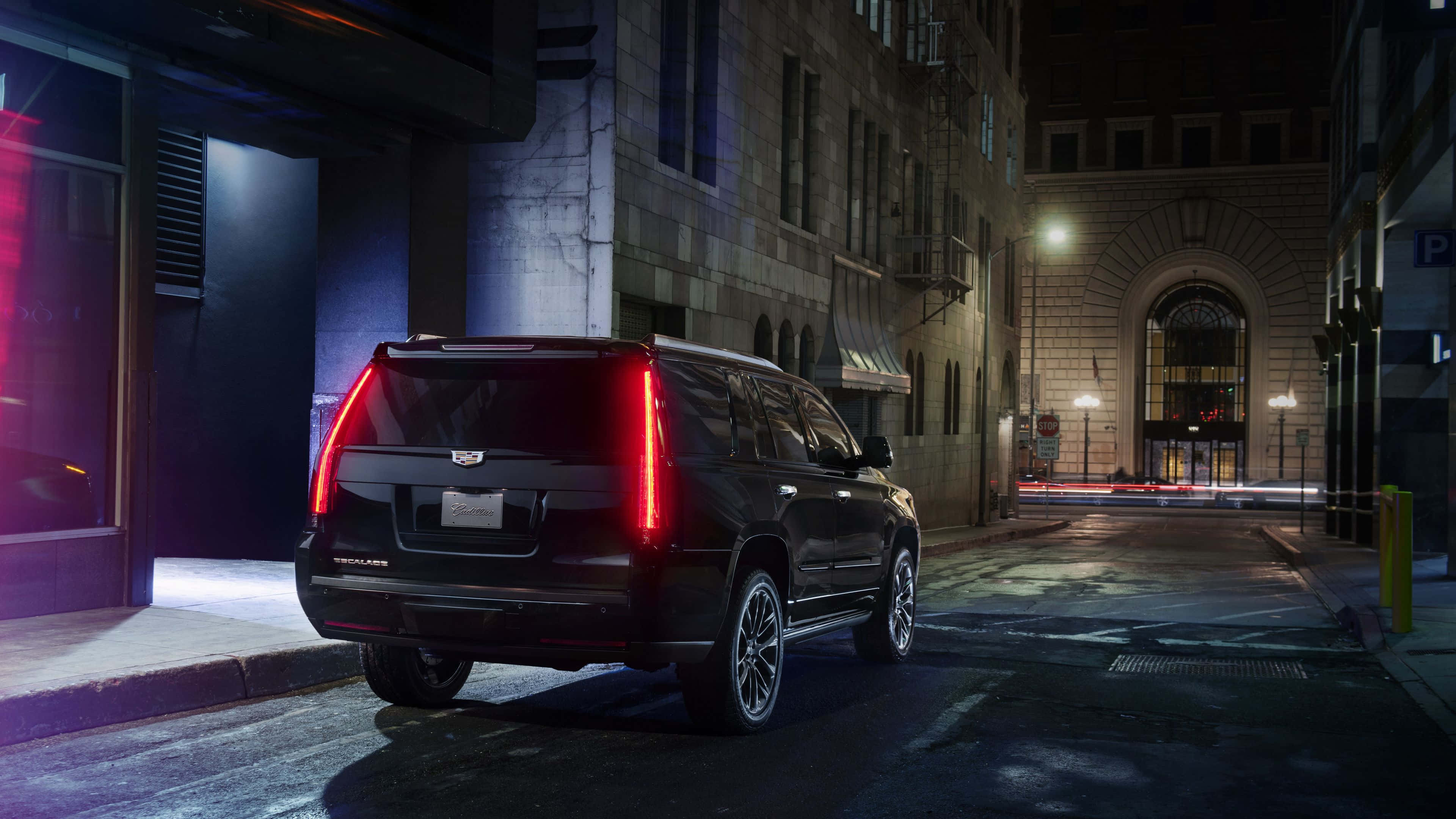 Cadillac Escalade 桌布 壁紙 — 一輛黑色Cadillac SUV在夜間停泊在濕漉漉的市區街道上，被霓虹燈和街燈照亮。