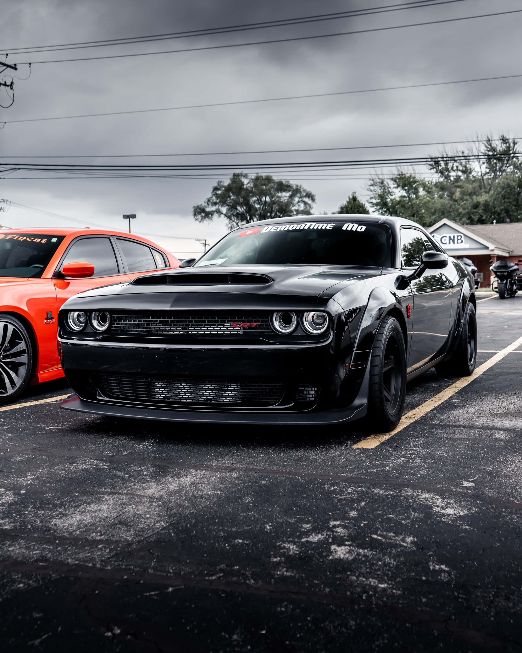 Dodge 桌布 壁紙 — 一輛黑色Dodge Challenger Hellcat帶有'DemonTime Mo'貼紙，停放在一個橙色汽車旁邊的車位，天空多雲。
