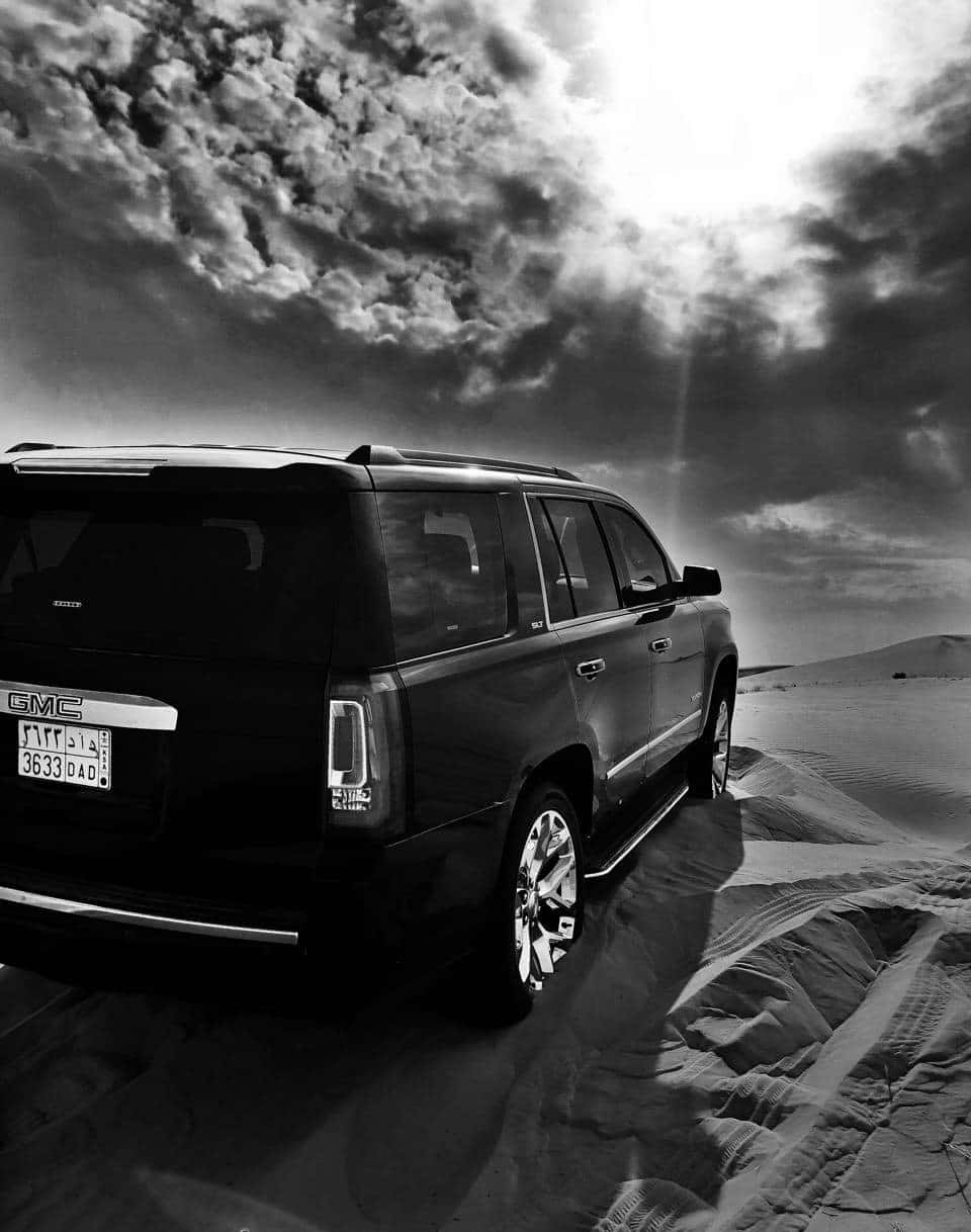 Gmc Yukon 桌布 壁紙 — 一輛黑色GMC SUV在單色調的戲劇性多雲天空下，矗立於紋理分明的沙漠沙丘之上。