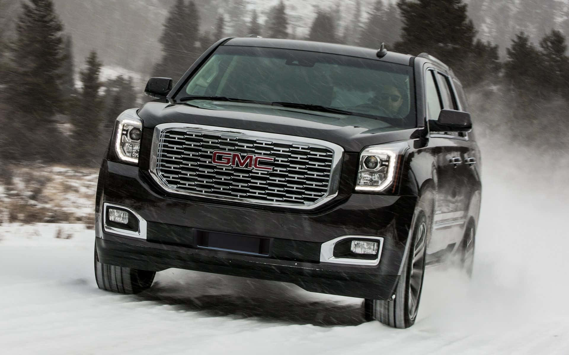 Gmc Yukon 桌布 壁紙 — 一輛黑色GMC SUV在深雪中行駛，輪胎濺起雪花，背景是覆蓋著白雪的常青樹。