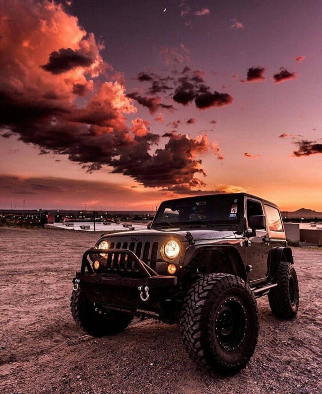 Wrangler 桌布 壁紙 — 一輛黑色Jeep Wrangler在碎石上，車頭燈閃爍，背景是壯麗的日落天空，帶有火紅色的橙雲。