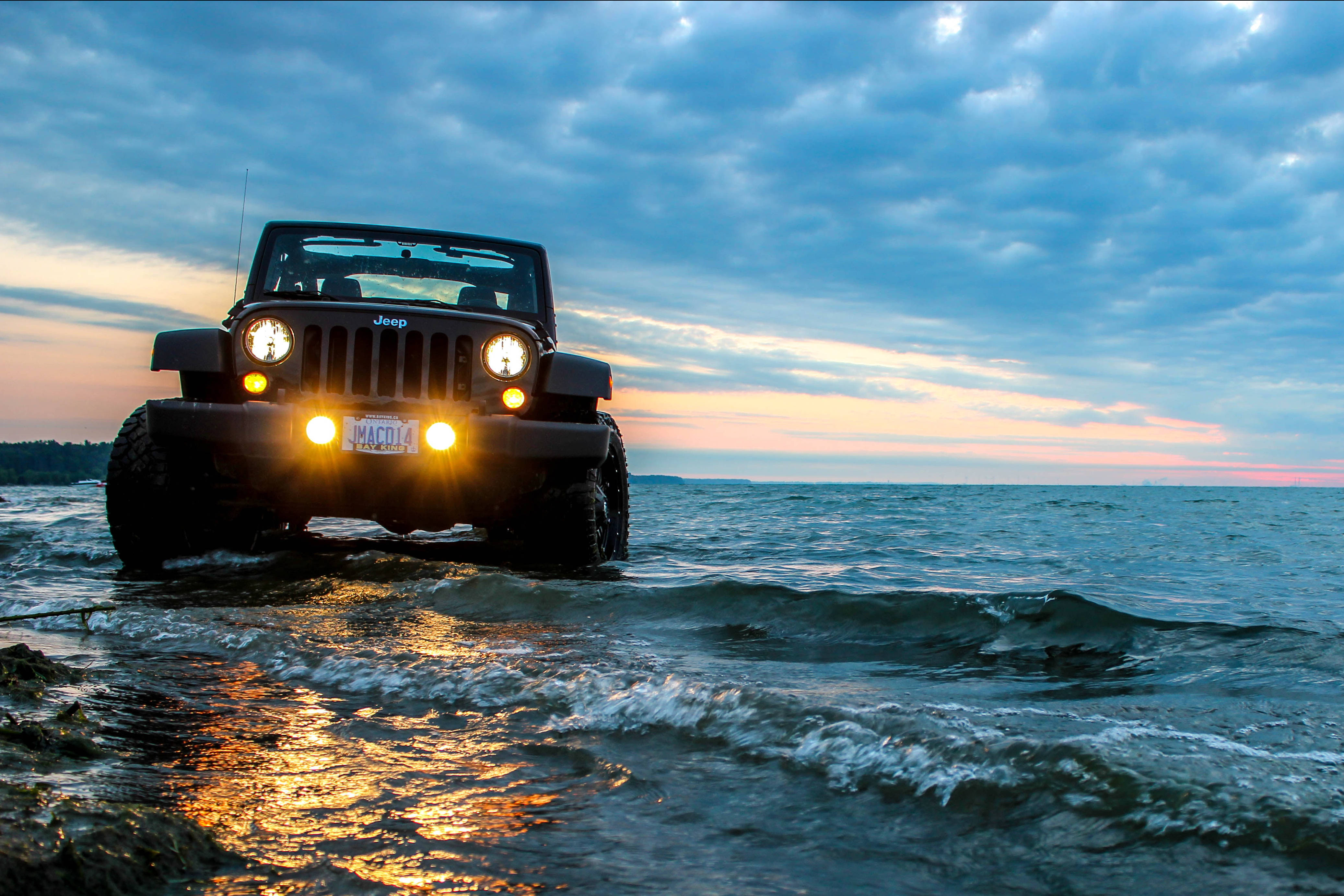 黑色吉普車 桌布 壁紙 — 一輛黑色Jeep Wrangler在日落時分涉水而過，明亮的車頭燈倒映在波浪上。