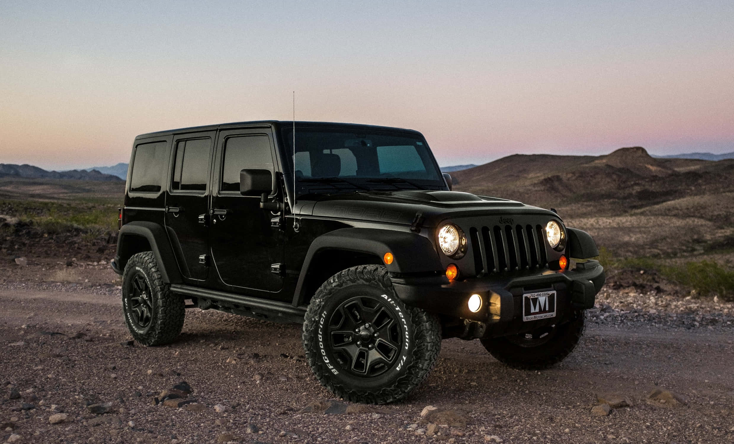 黑色吉普車 桌布 壁紙 — 一輛黑色Jeep Wrangler在岩石沙漠小徑上蓄勢待發，夕陽在遠山後落下，為天空染上溫暖色調。