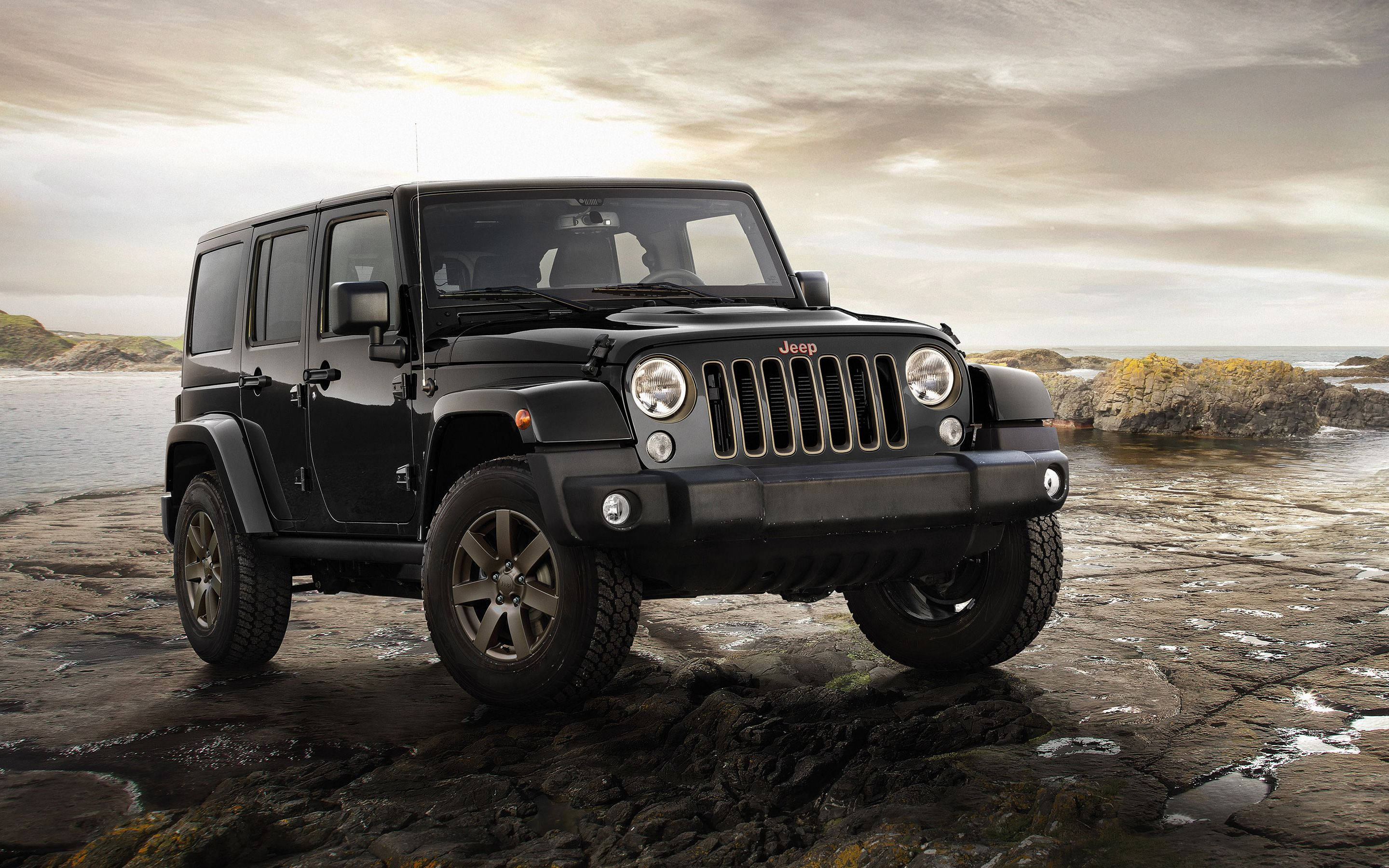 Wrangler 桌布 壁紙 — 一輛黑色Jeep Wrangler在陰天之下，堅固地停在濕漉漉的海岸岩石上，海浪在附近拍打。