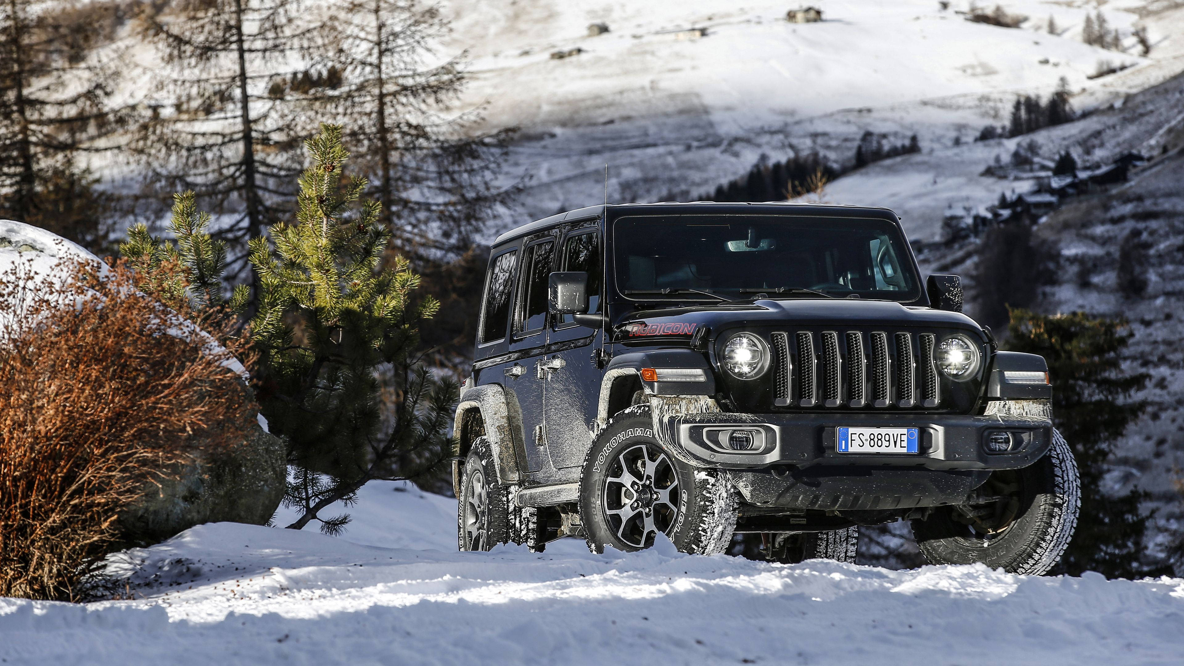 吉普 桌布 壁紙 — 一輛黑色Jeep Wrangler在明亮的日光下，穿越有松樹和岩石斜坡的雪山地形。