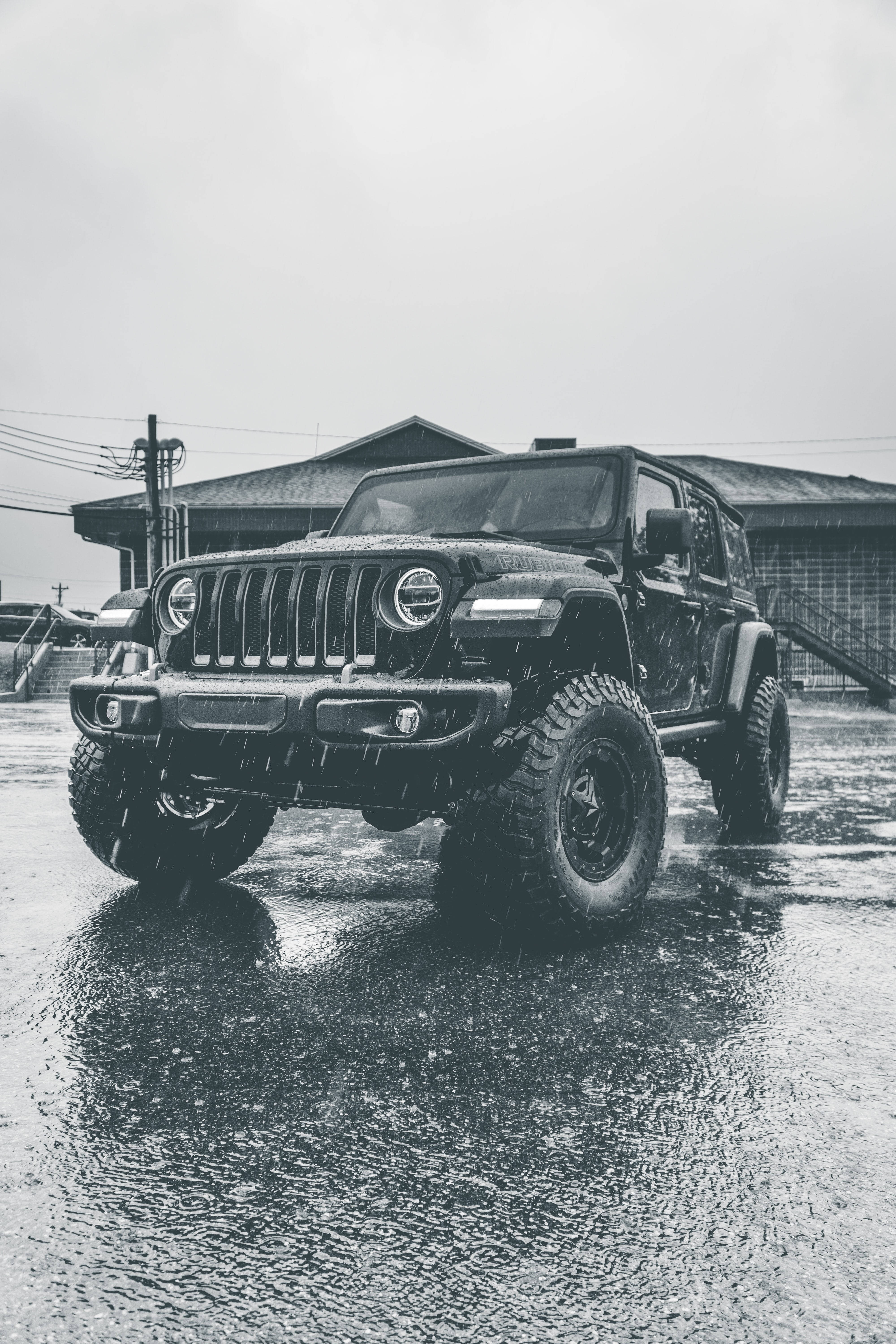 Black Jeep 桌布 壁紙 — 一輛黑色Jeep Wrangler在暴雨中堅毅地停在濕漉漉的瀝青停車場上，背景是一座建築物。